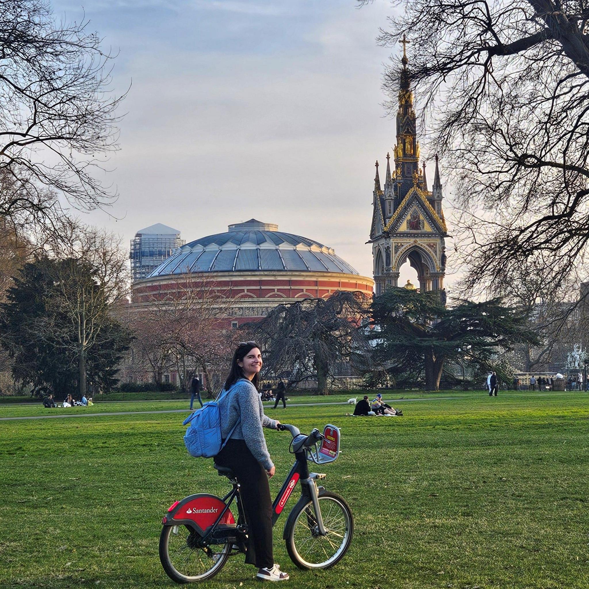 A woman with dark hair, wearing a grey jumper and carrying a blue rucksack, stands holding a Santander Cycle on the grass. The Royal Albert Hall and the Albert Memorial are behind her, with bare trees and people sitting in the park.