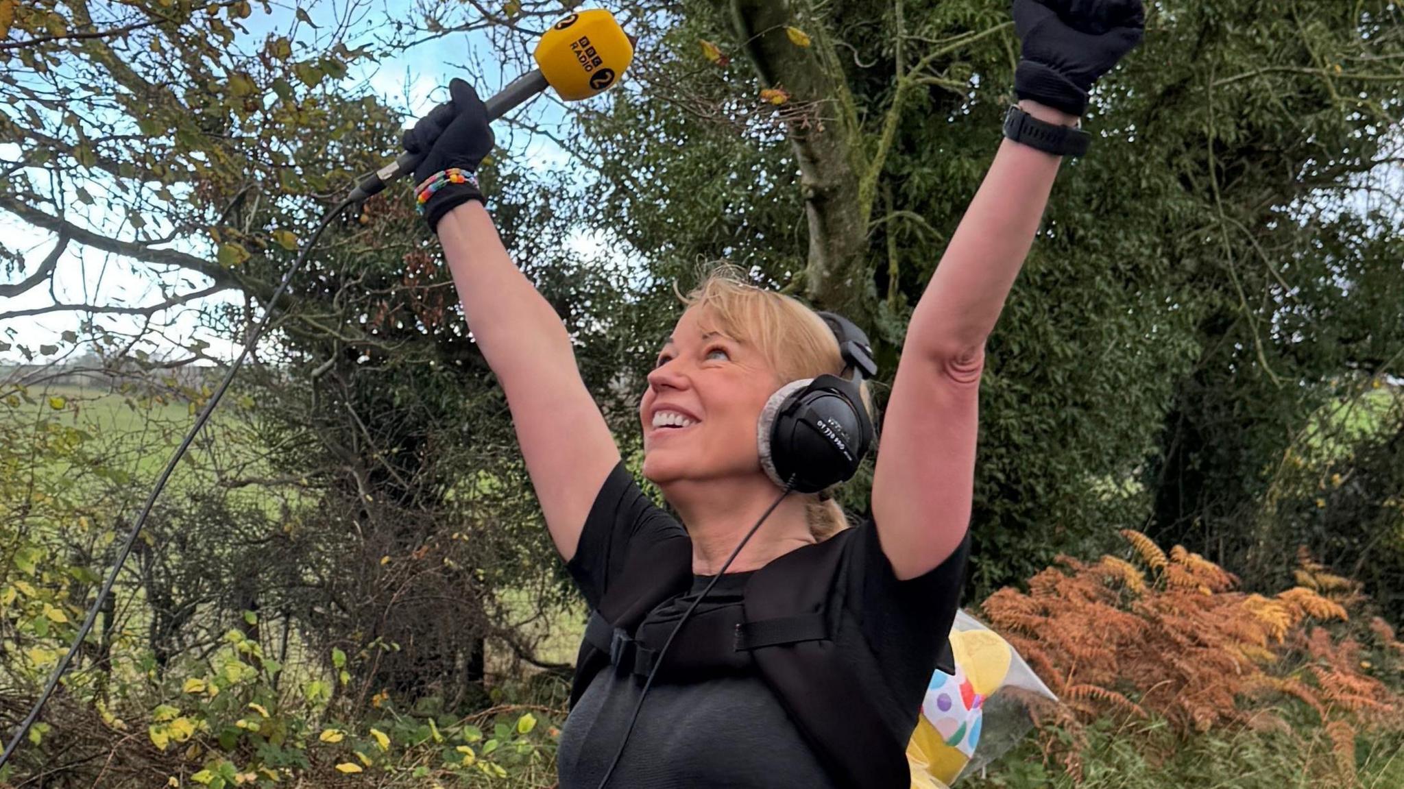 Sara Cox smiling with her hands in the air, she's wearing headphones and has a Radio 2 microphone in her hand