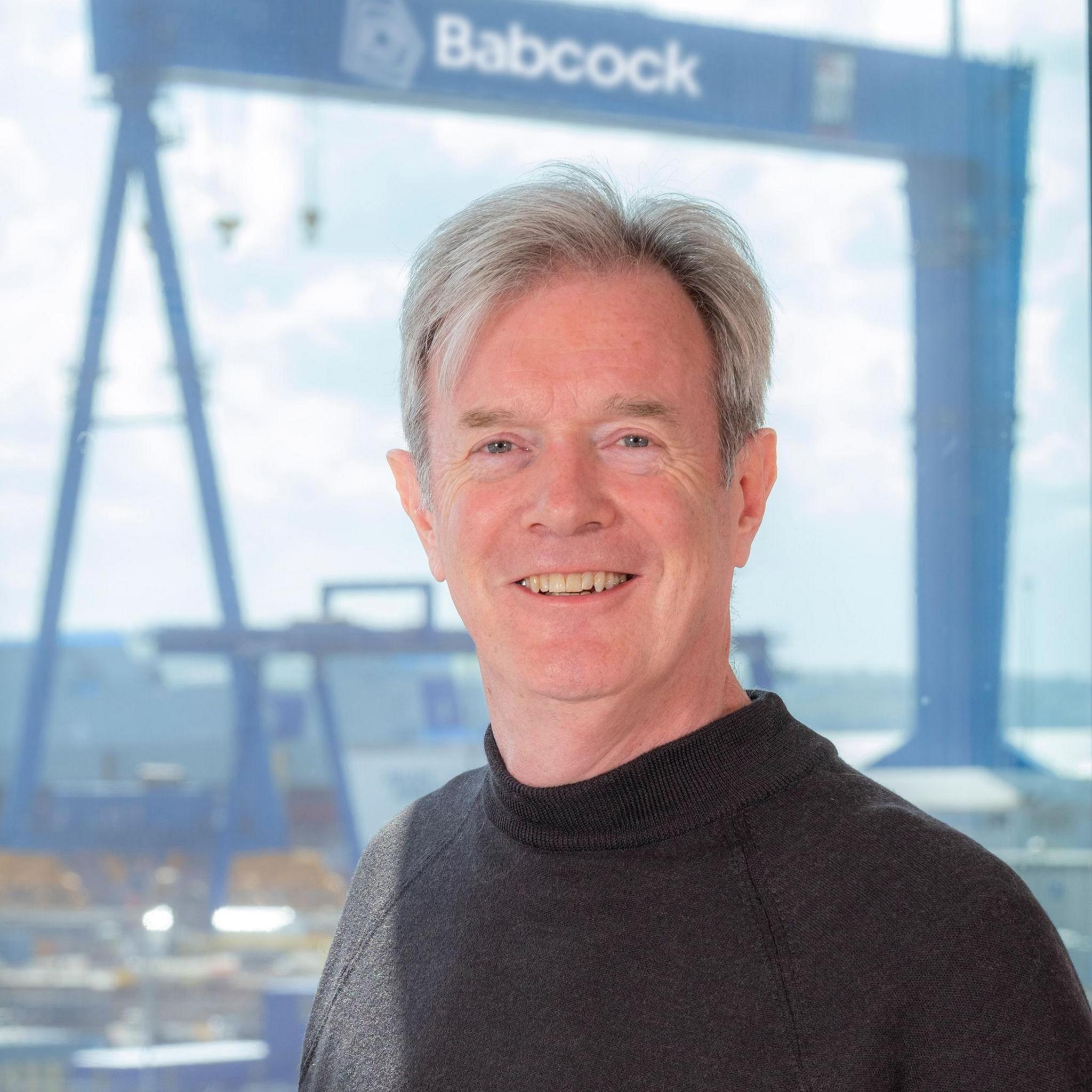 A man in a black sweater standing in front of a gantry crane with Babcock written on it