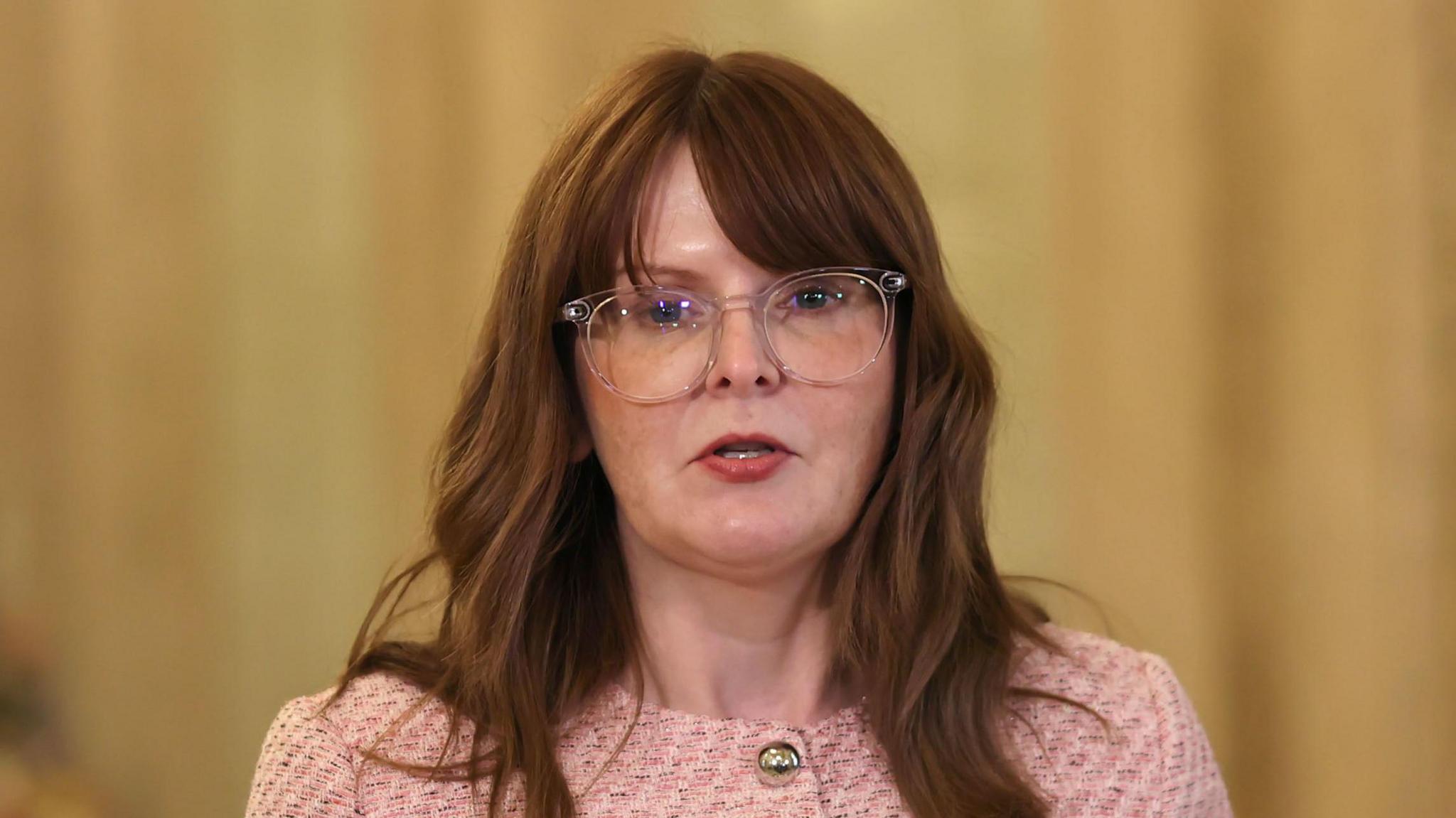 Caoimhe Archibald speaking into a microphone. She has long brown hair and is wearing a pink jacket with gold buttons and clear glasses.
