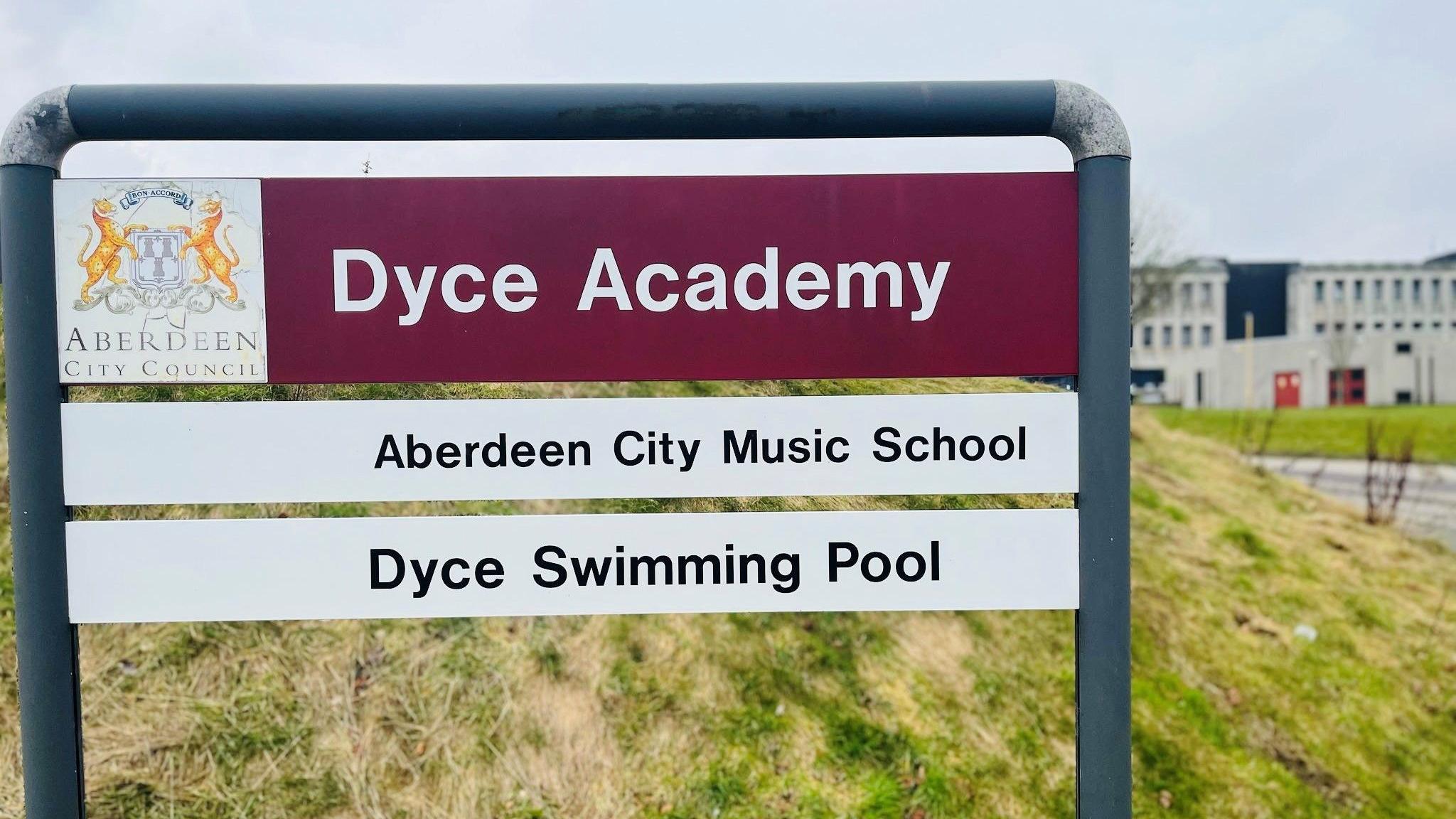 Aberdeen City Council sign which says 'Dyce Academy' in white on a burgundy strip, with school building in the background.