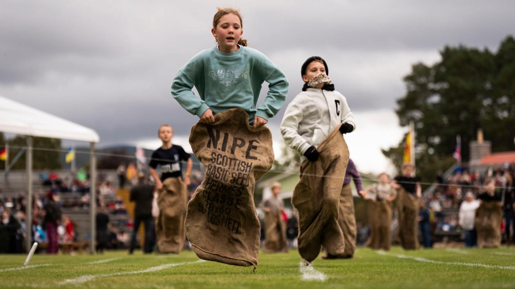 Children taking part in a sack race on a grassy field. Each participant is inside a large sack, hopping forward in the race. In the background, there's an audience observing the event