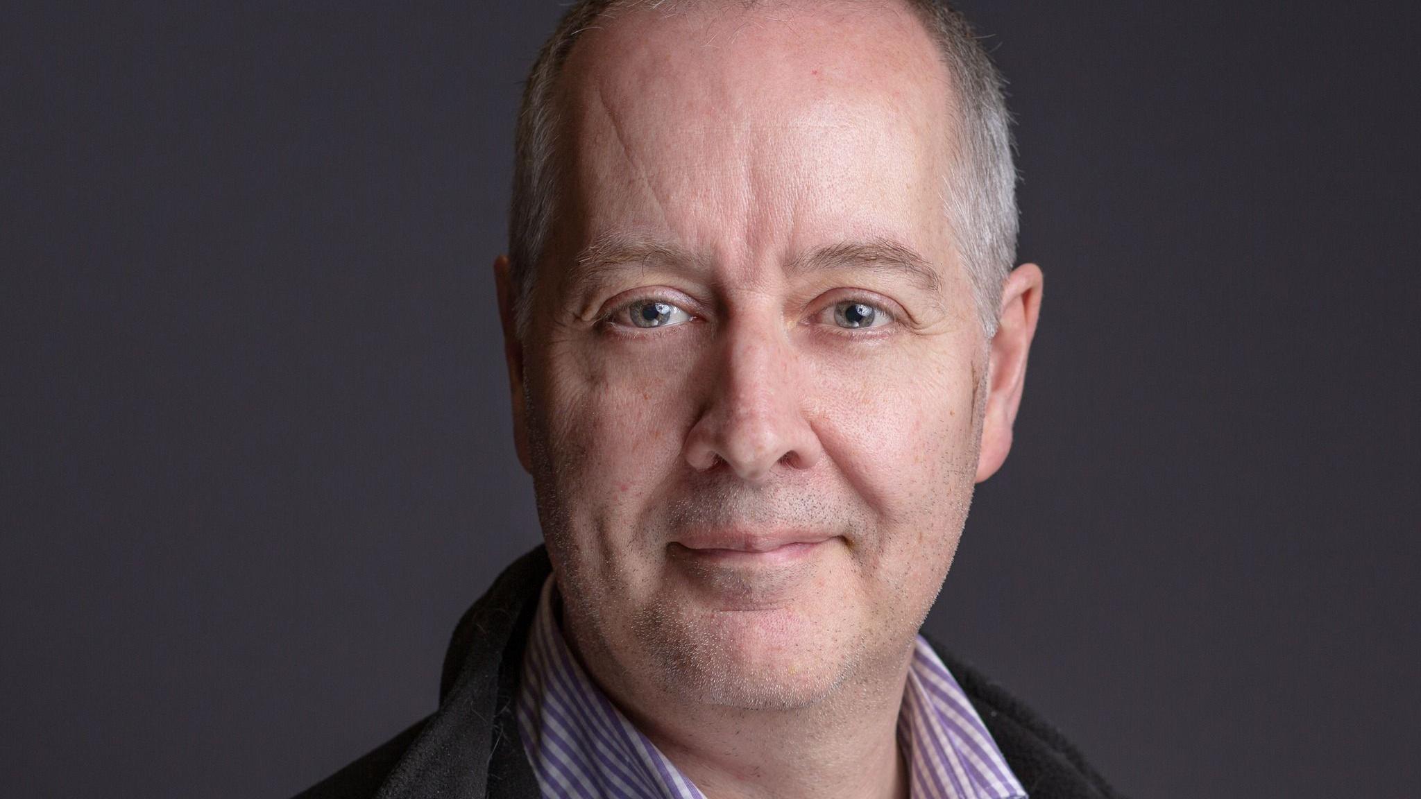 A council portrait of Simon Werner looking at the camera. He is wearing a stripy shirt and a green jacket 