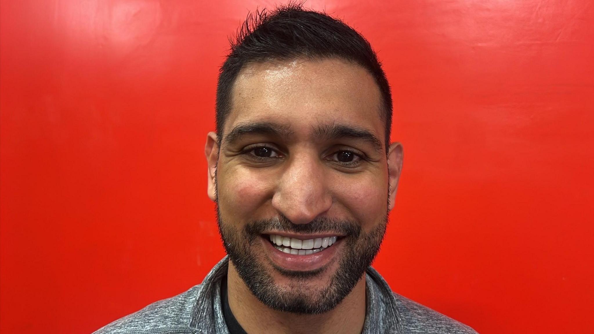 Amir Khan is looking towards the camera smiling. He has short black hair and brown eyes and is wearing a grey T-shit. He is stood in front of a bright red wall.