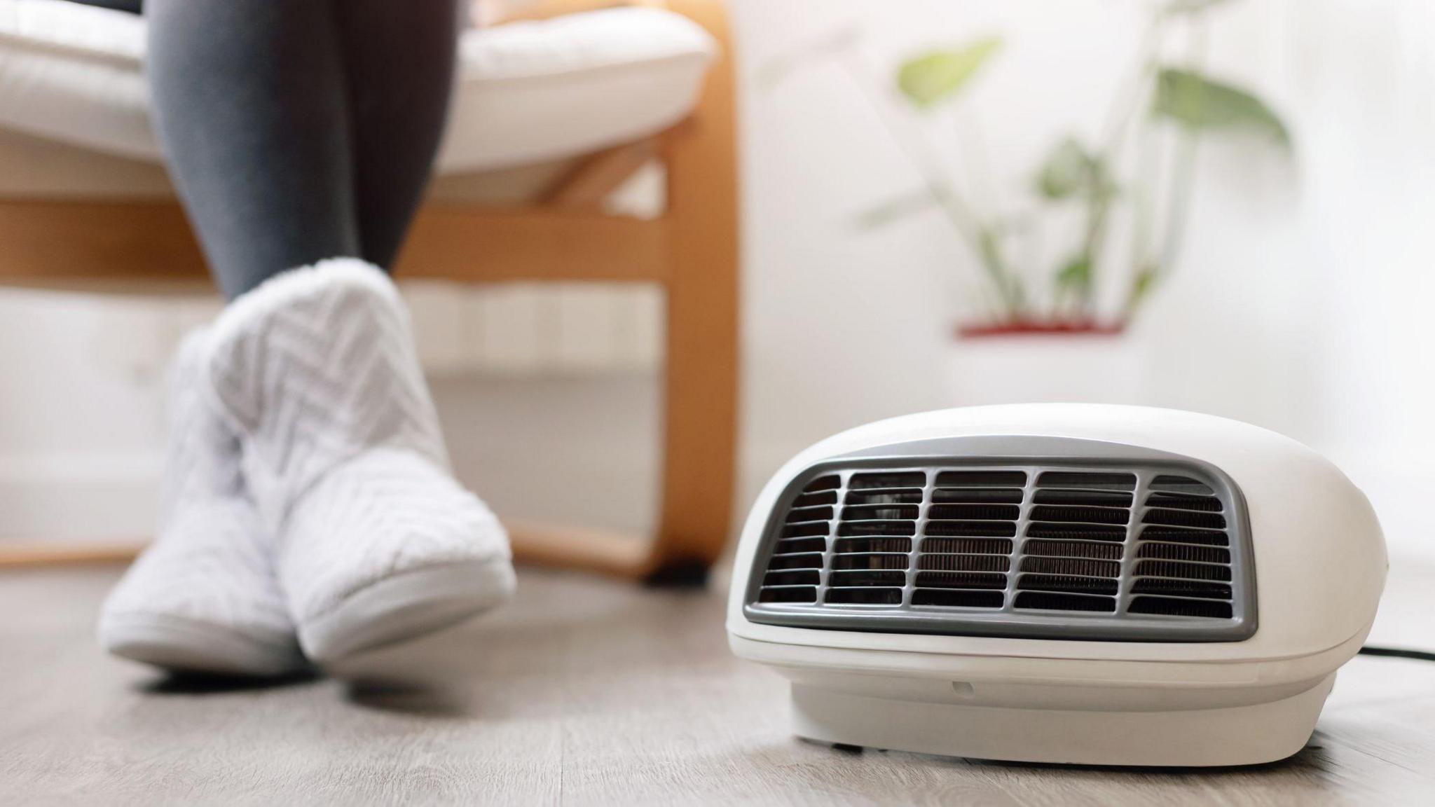 Close up shot of a woman wearing thick grey tights and white slipper boots next to an electric heater. 