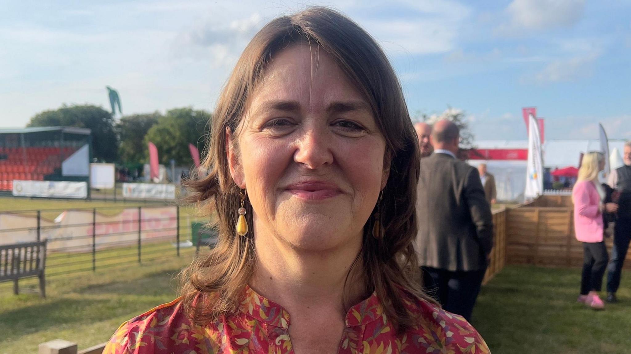 Julia Pyke is standing in a field with banners behind her, and is smiling looking towards the camera. She has shoulder length brown hair, and is wearing a pink patterned top.