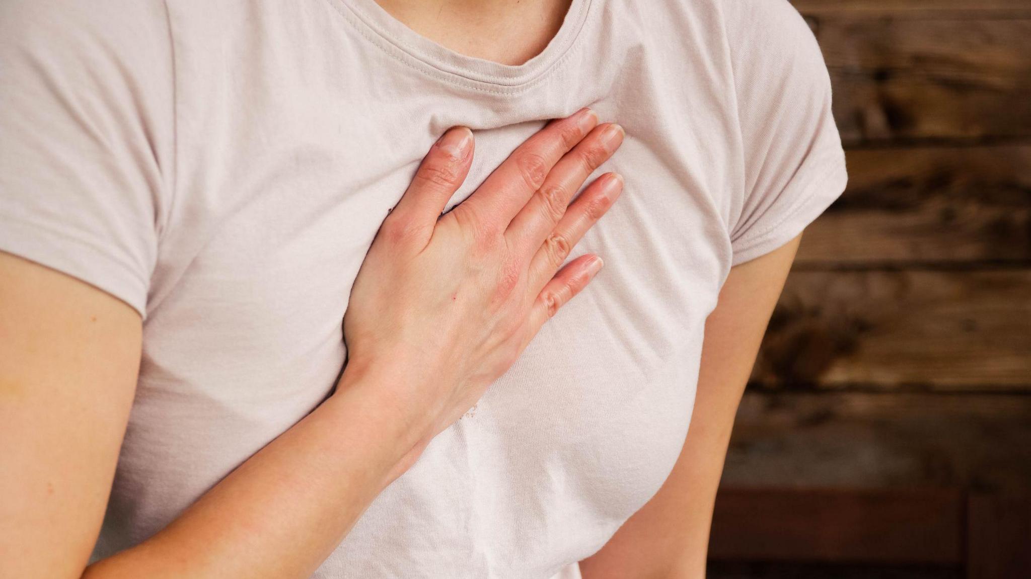 A woman is holding her hand on her chest to indicate a problem with her heart. She is wearing a light pink T-shirt