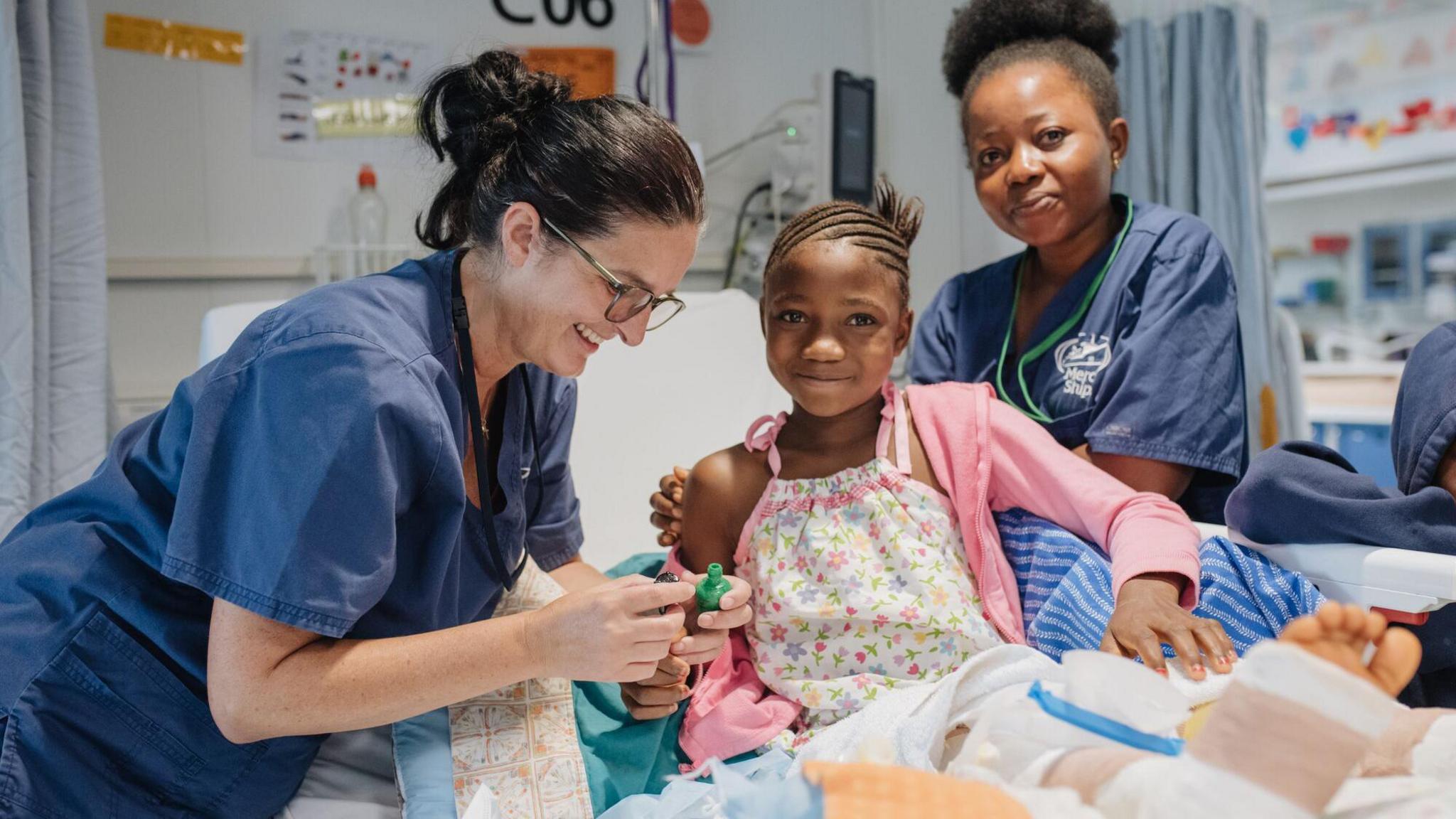 Fudia is in a hospital bed, smiling at the camera, she is sat upright, her hair in braids wearing a white nightie with pink and yellow flowers and a pink unzipped hoodie over the top. Dr Rachel Buckingham is standing to Fudia's left, she has black hair tied up in a bun and wears plastic framed glasses and navy blue scrubs, she is smiling while looking down at a syringe tending to Fudia. A second medic is standing behind Fudia, wearing navy blue scrubs, her hair tied up in a high ponytail and smiling at the camera