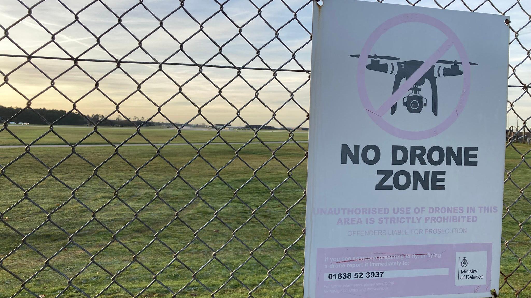 Sign prohibiting drones attached to the fence of RAF Lakenheath in Suffolk. Behind the fence is grass and a runway. The sign has an image of a drone.