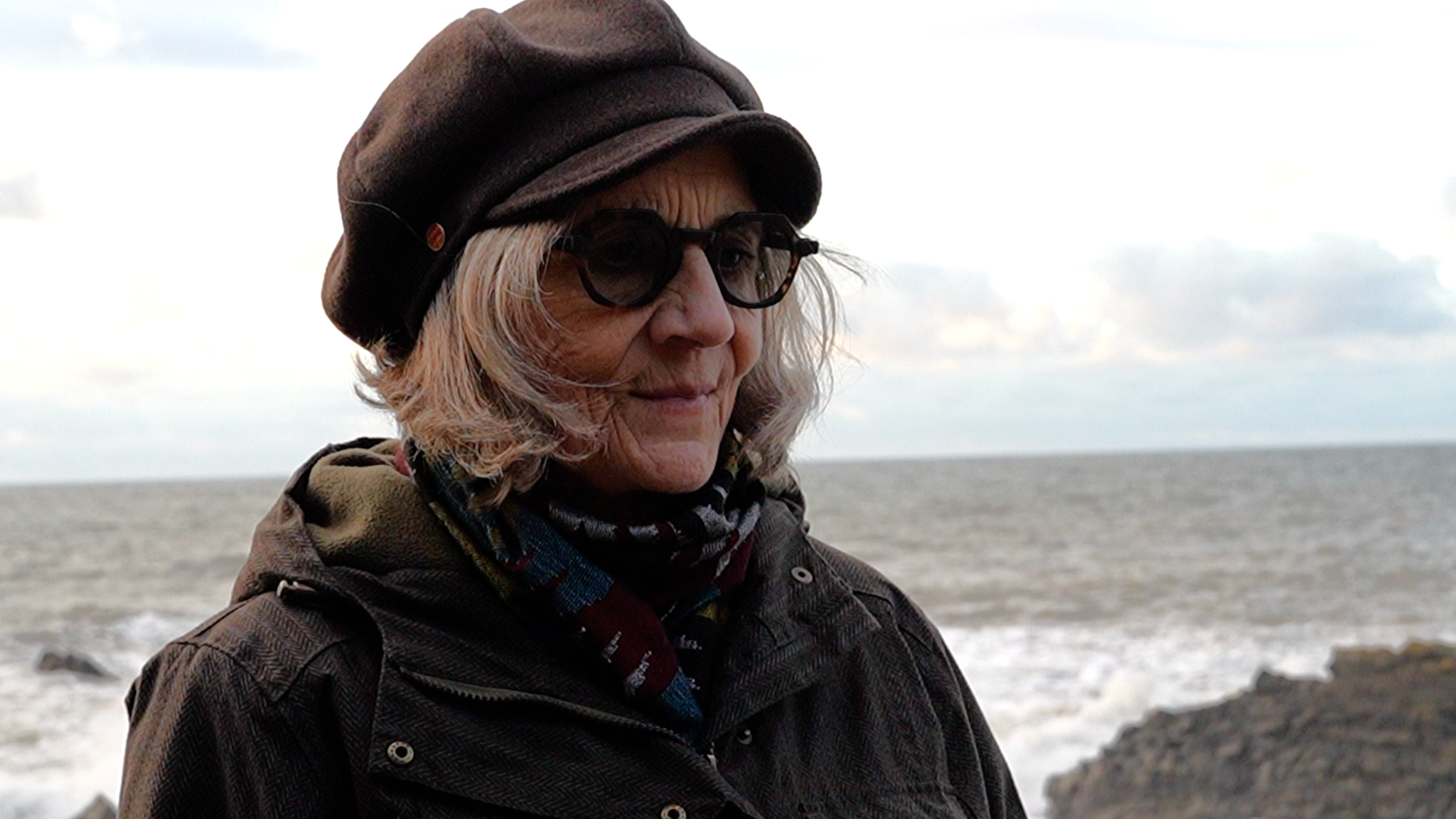 Carolyn Challis stood on a rocky coastline on a grey overcast day. She is wearing a brown coat, a scarf and a hat. Behind her the sea looks grey and choppy as waves hit the rocks.