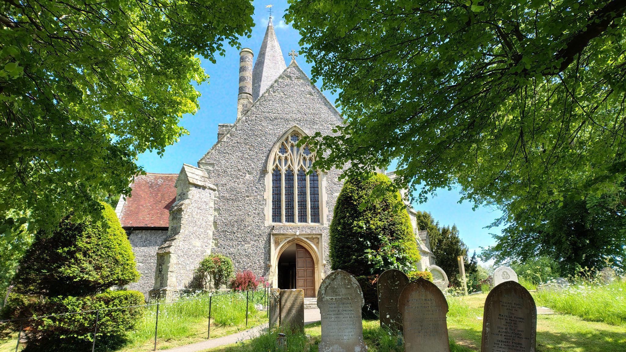 The exterior of St Andrew's Church in Alfriston, a 14th century building with a steeple and several graves in the foreground.
