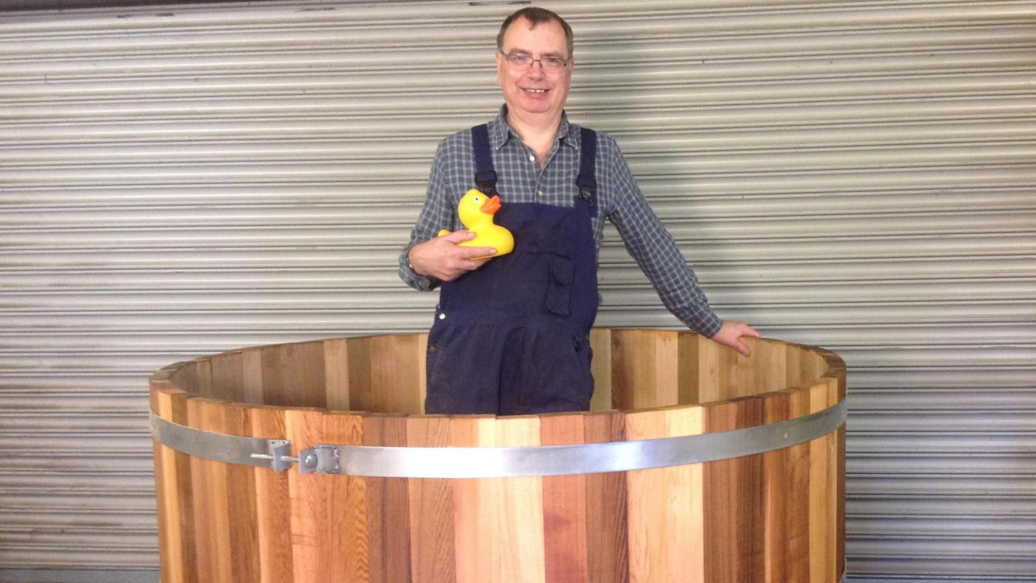 Alan Paulus wearing building wear and standing in a large wooden hot tub, while holding a rubber duck.