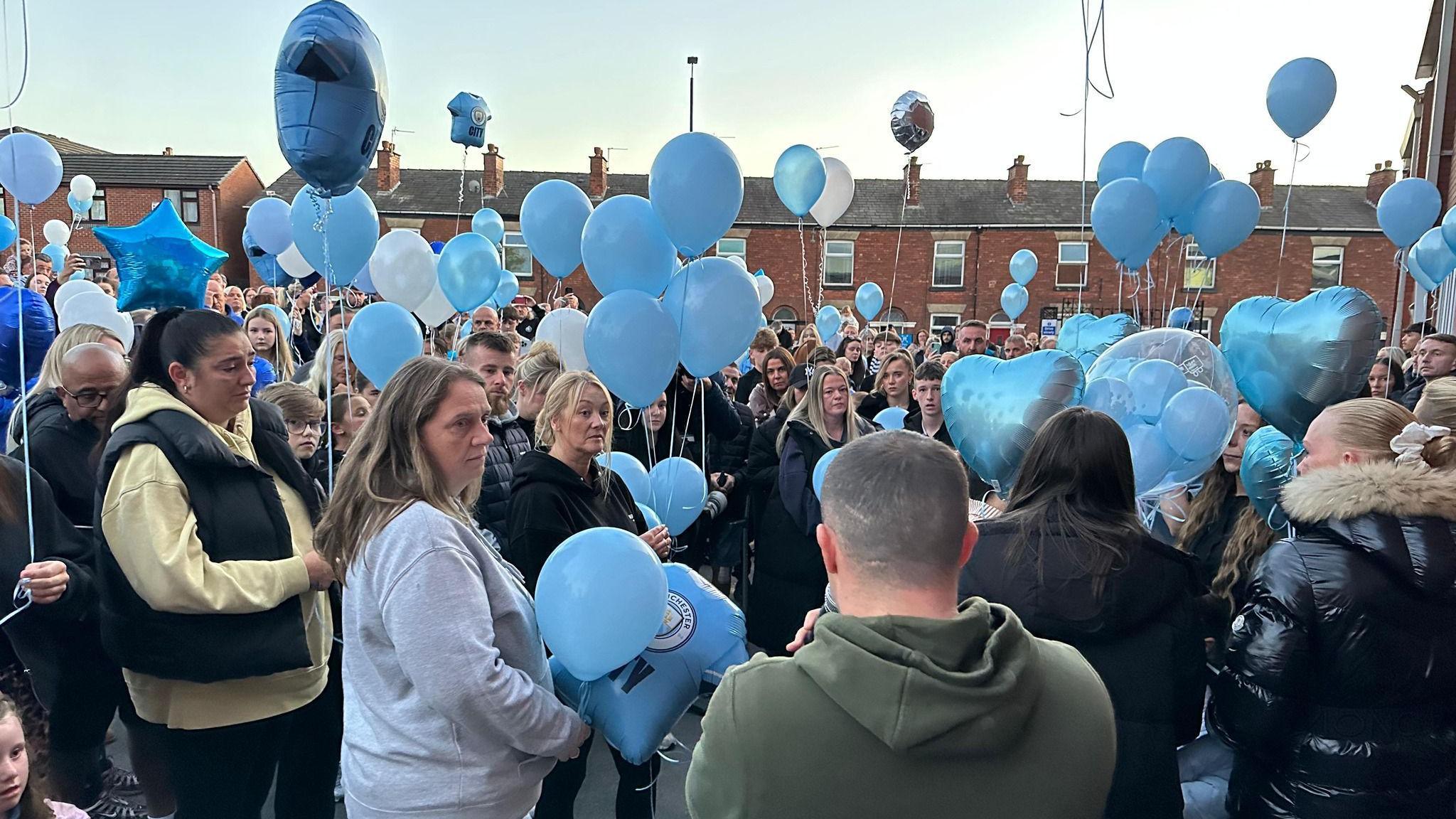 A large crowd of people stands in a street surrounded by brick houses and holding light blue balloons. A man in the foreground is seen facing away from the crowd and speaking into a microphone. 
