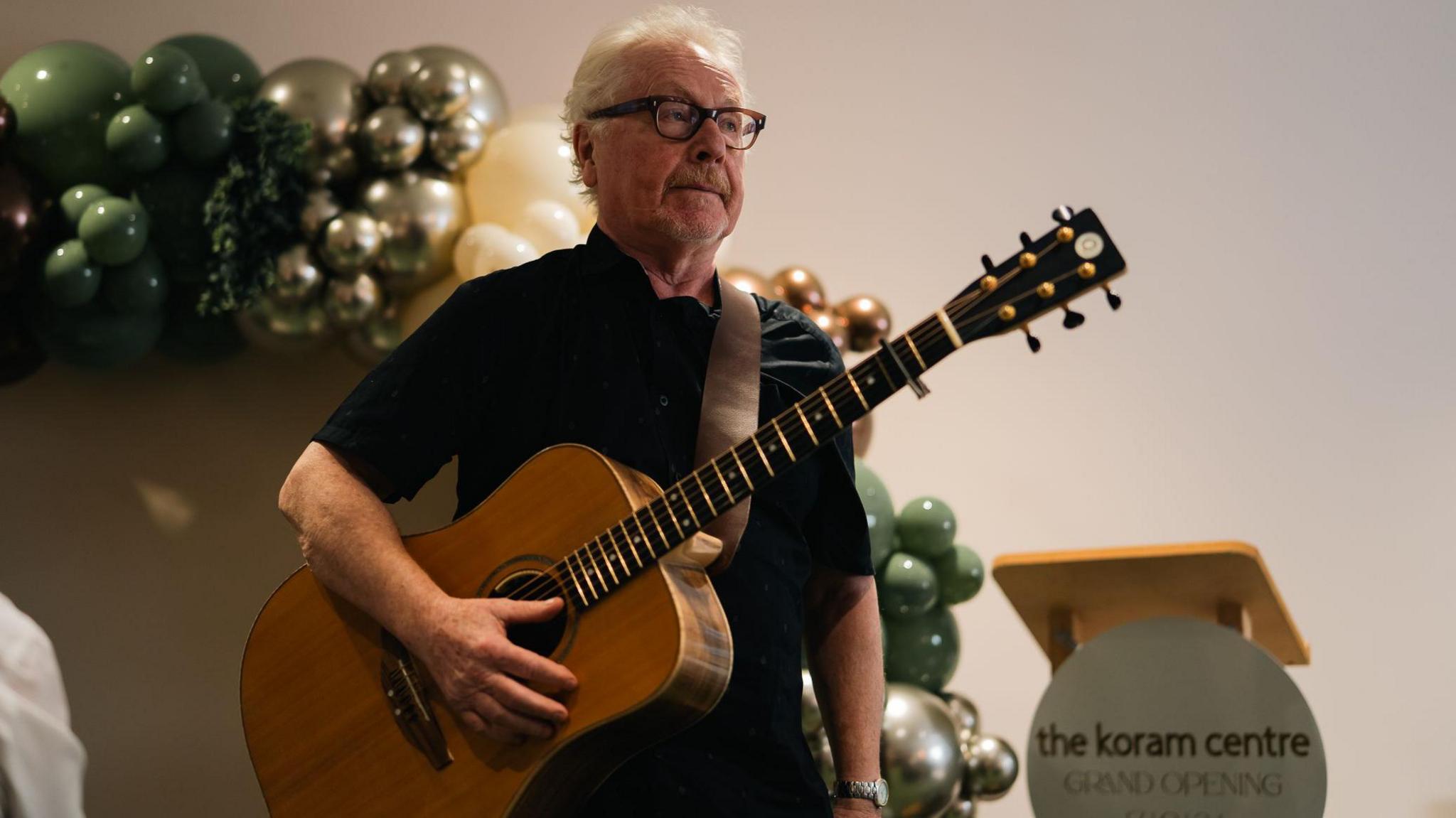 Paul brady holding a guitar. He is wearing a black t-shirt and glasses and has white hair. There is a balloon arch on the left and a podium on the right with a sign saying 'the koram centre GRAND OPENING'