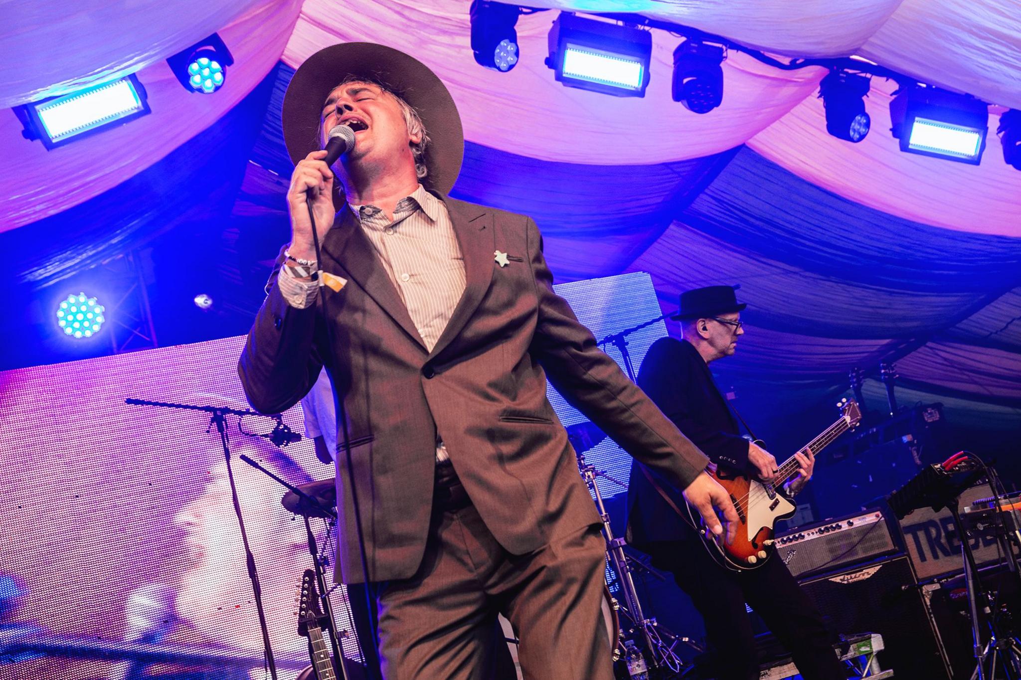 Singer Pete Doherty wears a wide-brimmed hat, and dark brown suit with light brown shirt.  He is singing into the microphone with a guitarist in the background. 