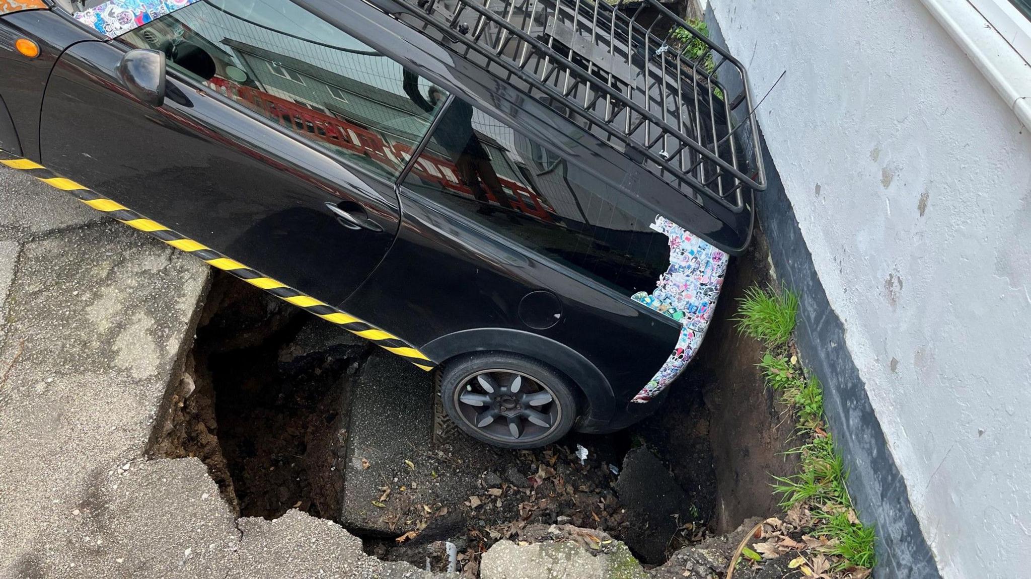 A Mini car's rear has fallen into a hole around 2 metres deep. Metal fencing has been put up around the area which is a car park behind a bakery in Redruth.