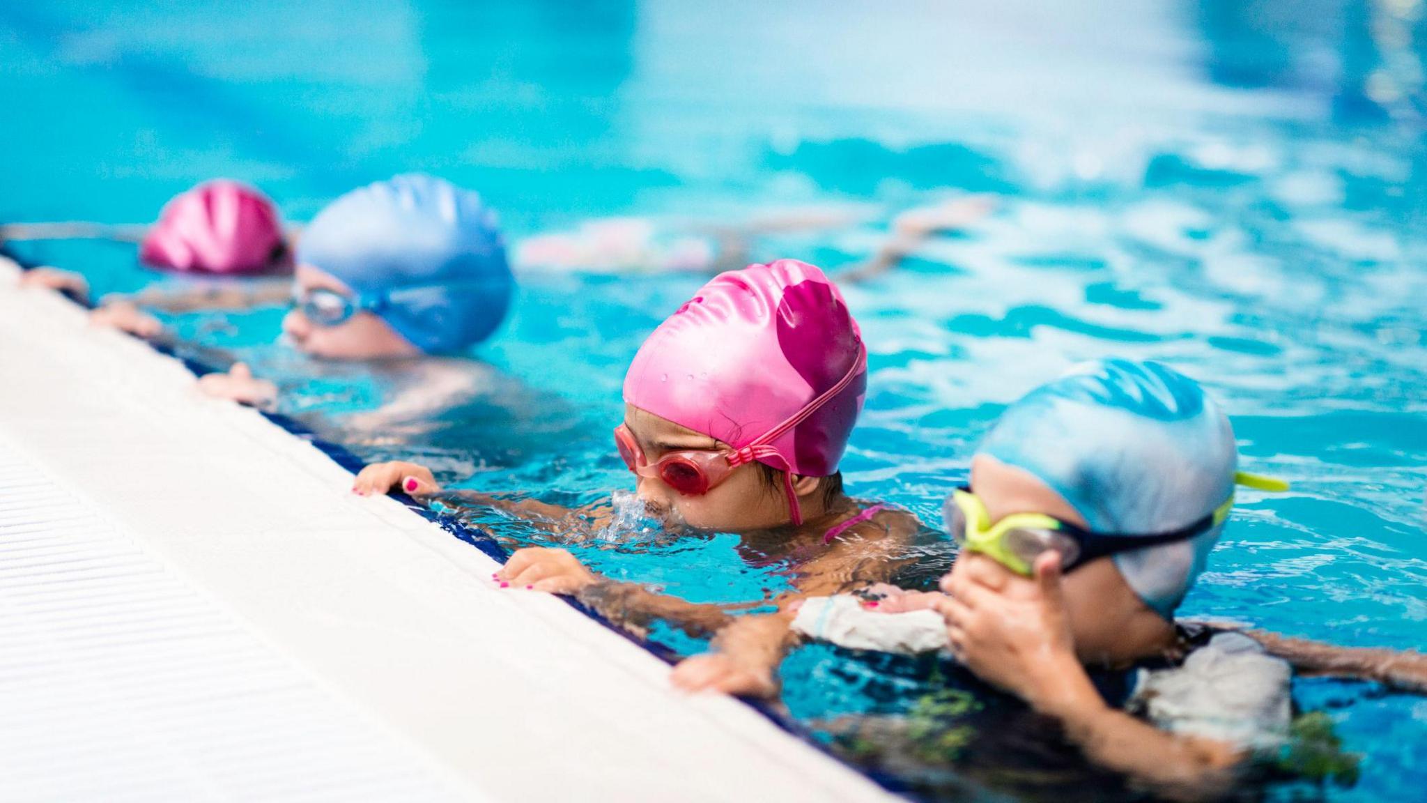 Five children in swimming caps and goggles are lined up along the edge of a bright blue swimming pool, learning to swim. The child closest to the camera wears a light blue cap and yellow goggles, with a second child in a pink cap and red goggles just behind them.