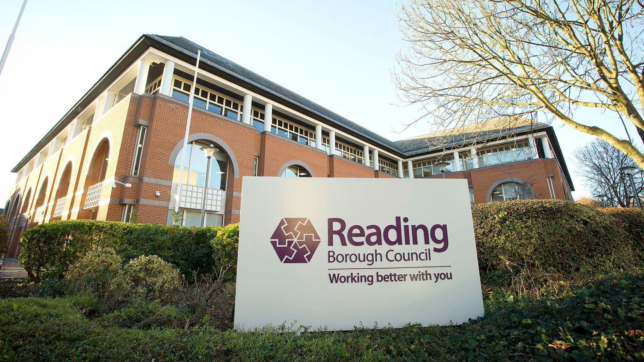 A brick building with a hedge outside, with a large white and purple sign saying 'Reading Borough Council, working better with you'.