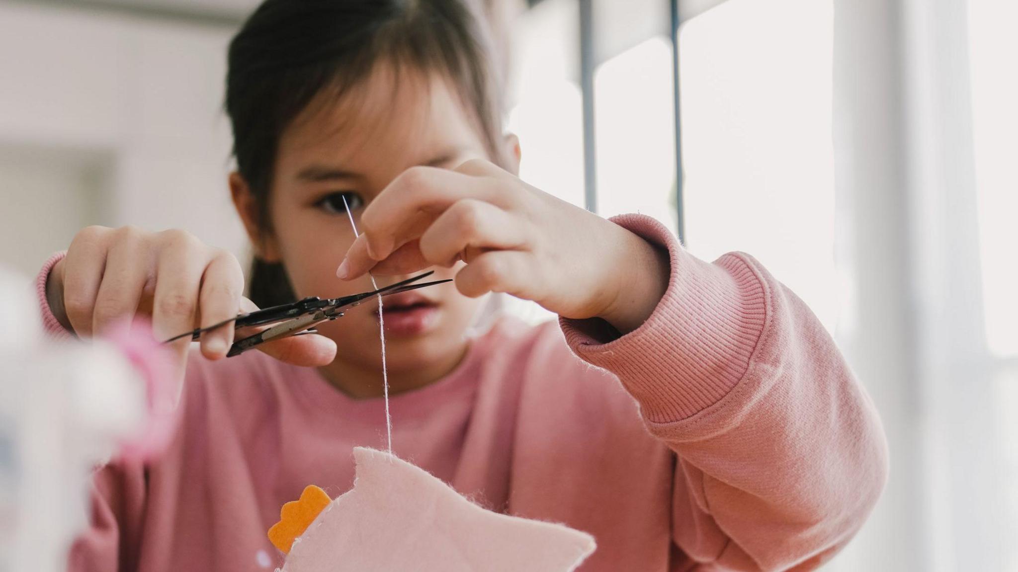 A young girl wearing a pink sweatshirt sewing felt at home