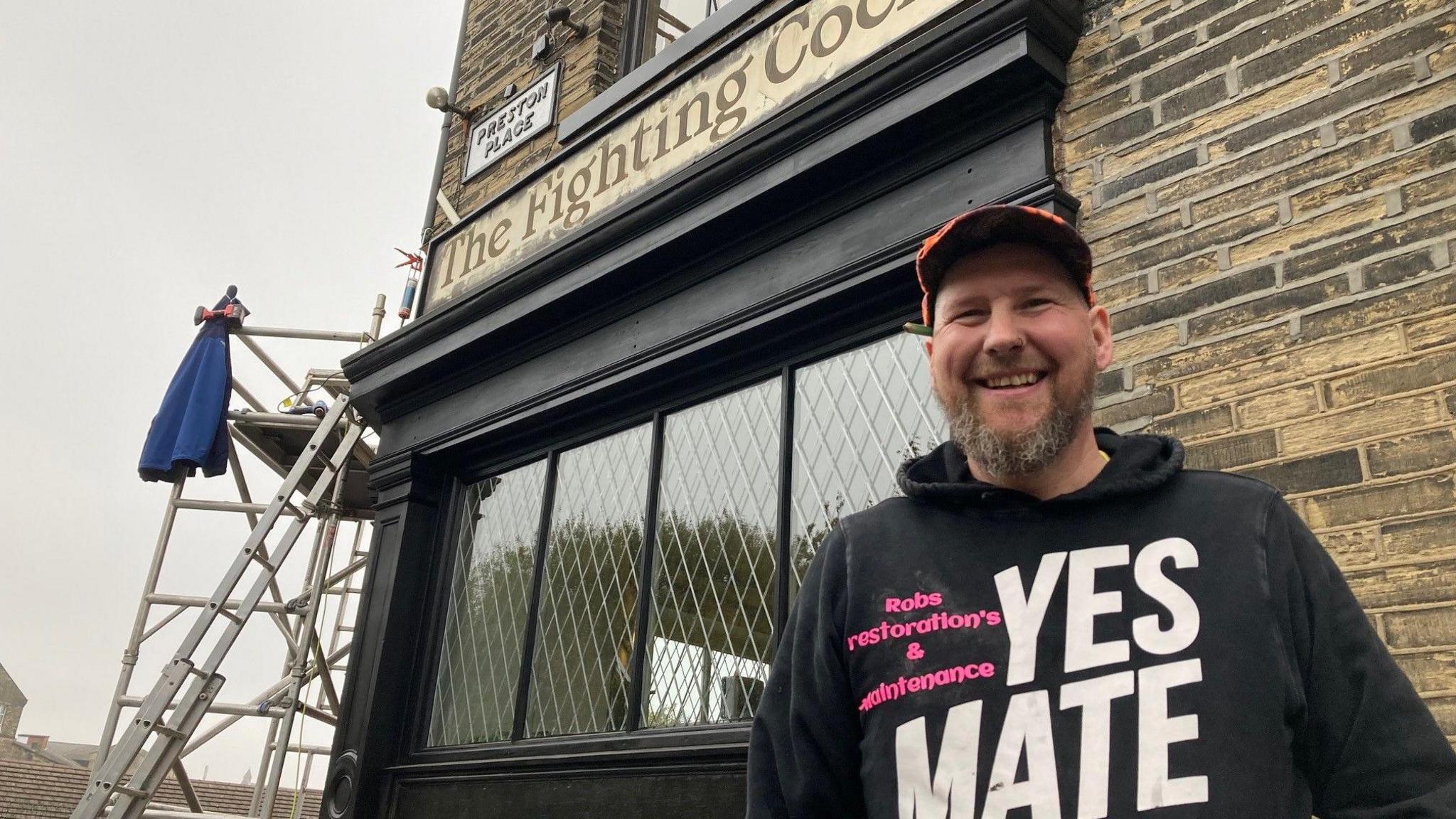 A man in an orange peaked-cap with a pencil tucked in behind one of his ears and wearing a black sweatshirt with white writing spelling out 'Yes mate' and also Rob's Restorations and Maintenance in smaller pink letters with the pub exterior in the background. 