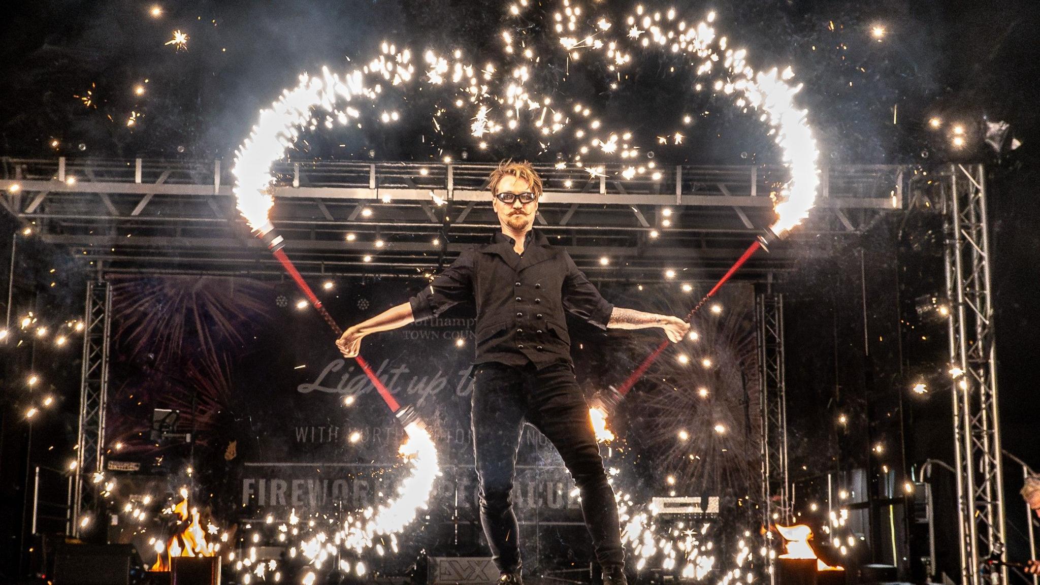 A person holding batons which have sparklers coming out the end of them. He is wearing all black and is stood on a stage at a fireworks display event.