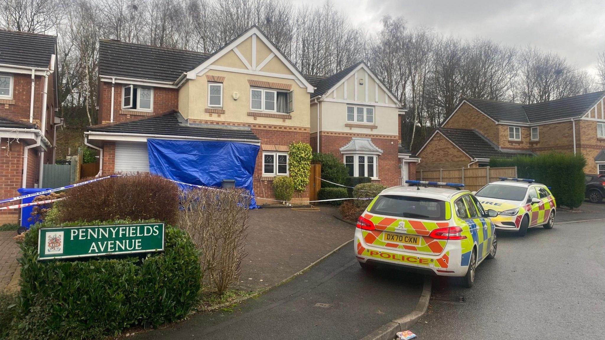 A police car and a fire service vehicle are parked outside a property where the windows of the top floor are open and there is blue tarpaulin covering part of the lower frontage of the house.