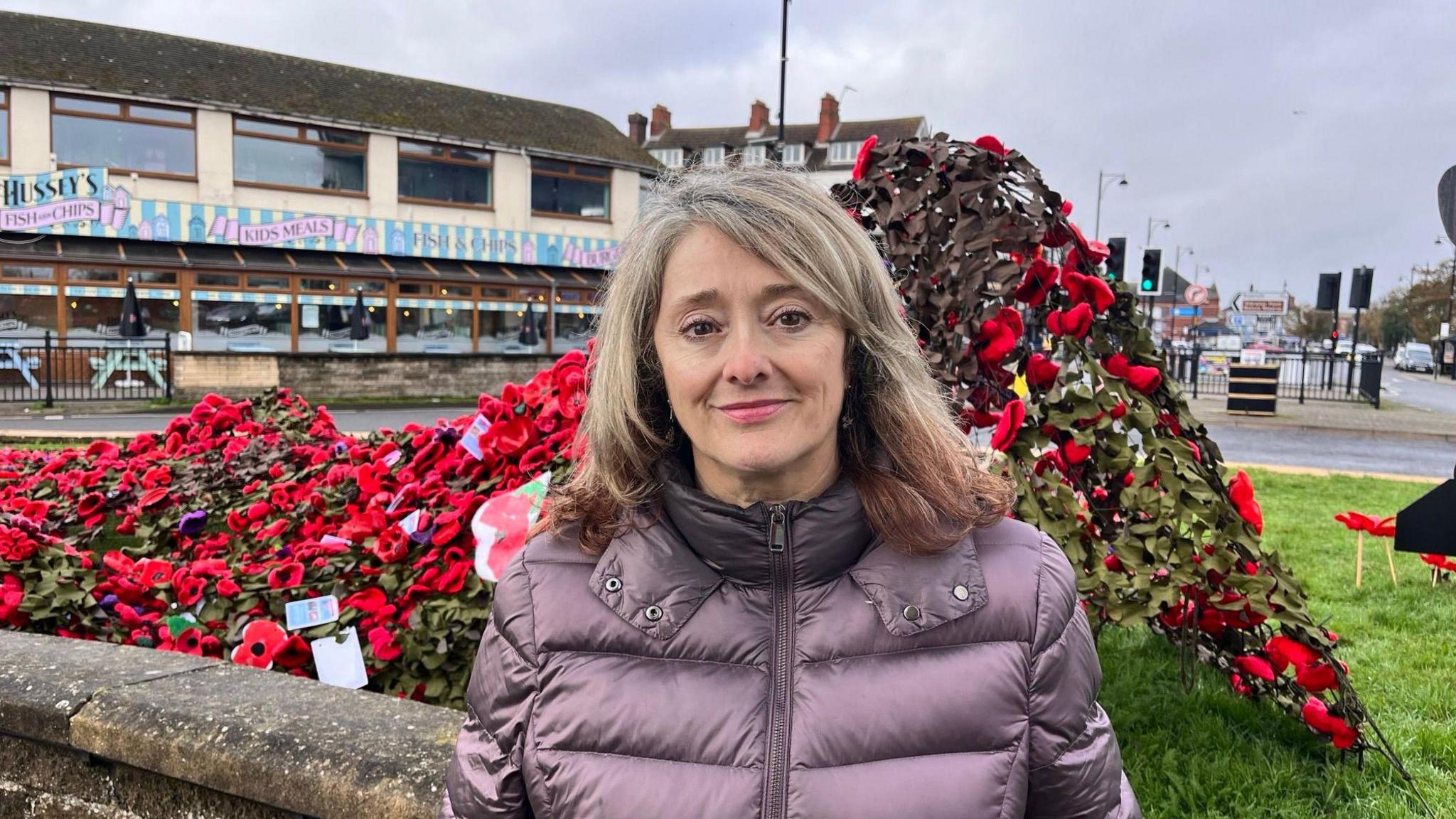 A woman with blonde and brown ombre hair wearing a purple coat. She is stood in front of a poppy display. 