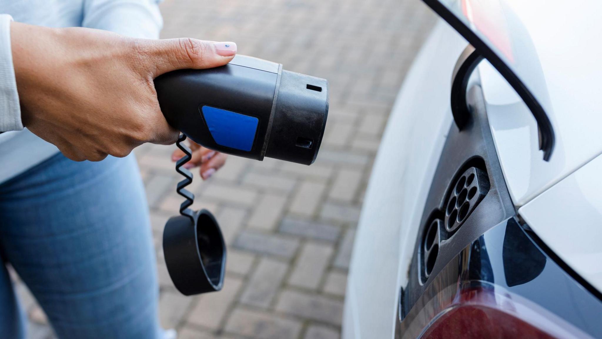 A woman prepares to slot an electric vehicle charger into a port on a car. The cap for the front of the charger dangles by a cord.