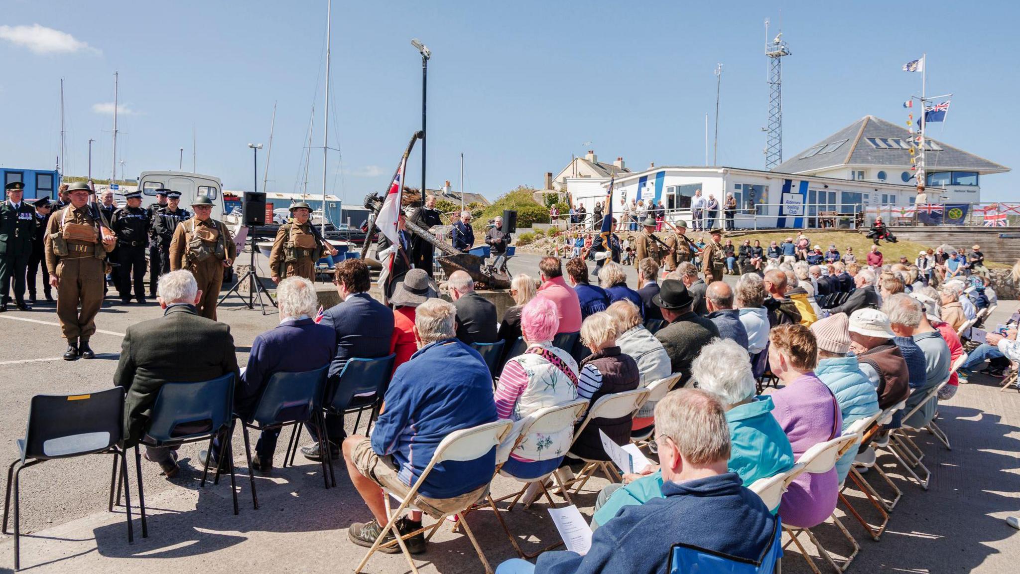 A large crowd of seated islanders are seen watching a speech. The speech maker is stood in front of a plinth with soldiers in historic military dress either side of them. 