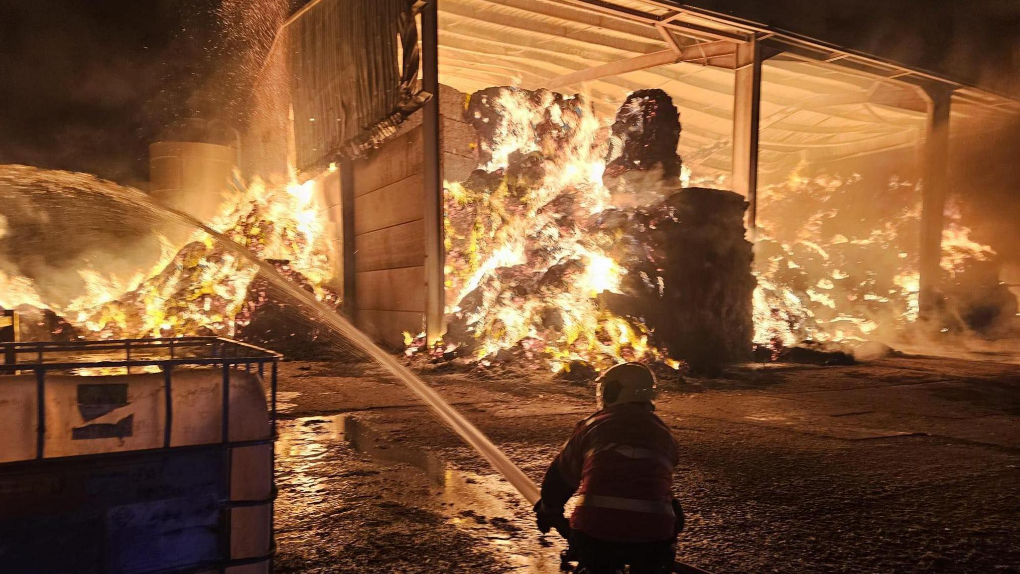 Straw in a barn is burning brightly int he dark as a firefighter uses a hose to combat the flames.