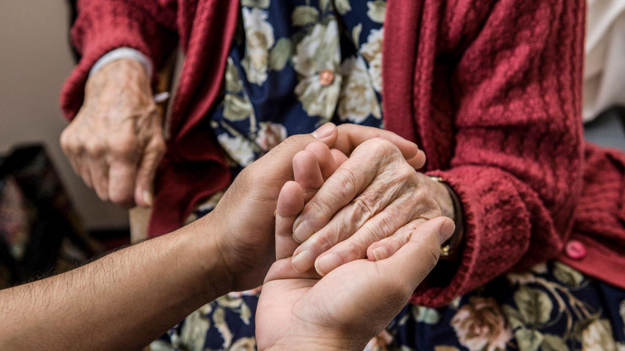 An elderly person in a red cardigan and floral dress has their hands gently held by another individual.