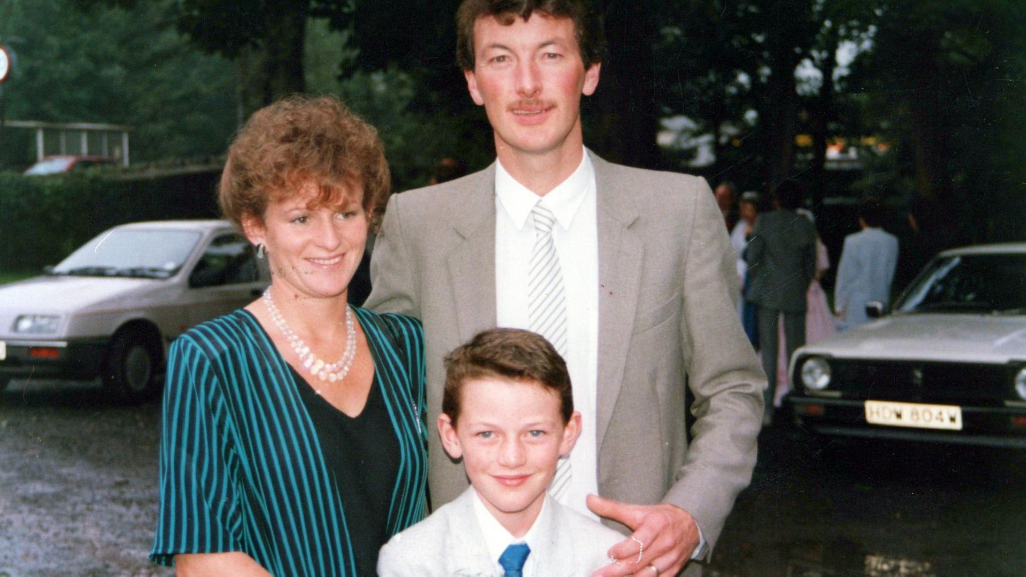 Luke Evans as a young boy with his parents Yvonne and David. Luke is in a pale grey suit and blue tie. David is in a grey suit, white shirt and striped tie on the right, and Yvonne is in a blue and black pinstriped jacket and black top, with pearls around her neck.