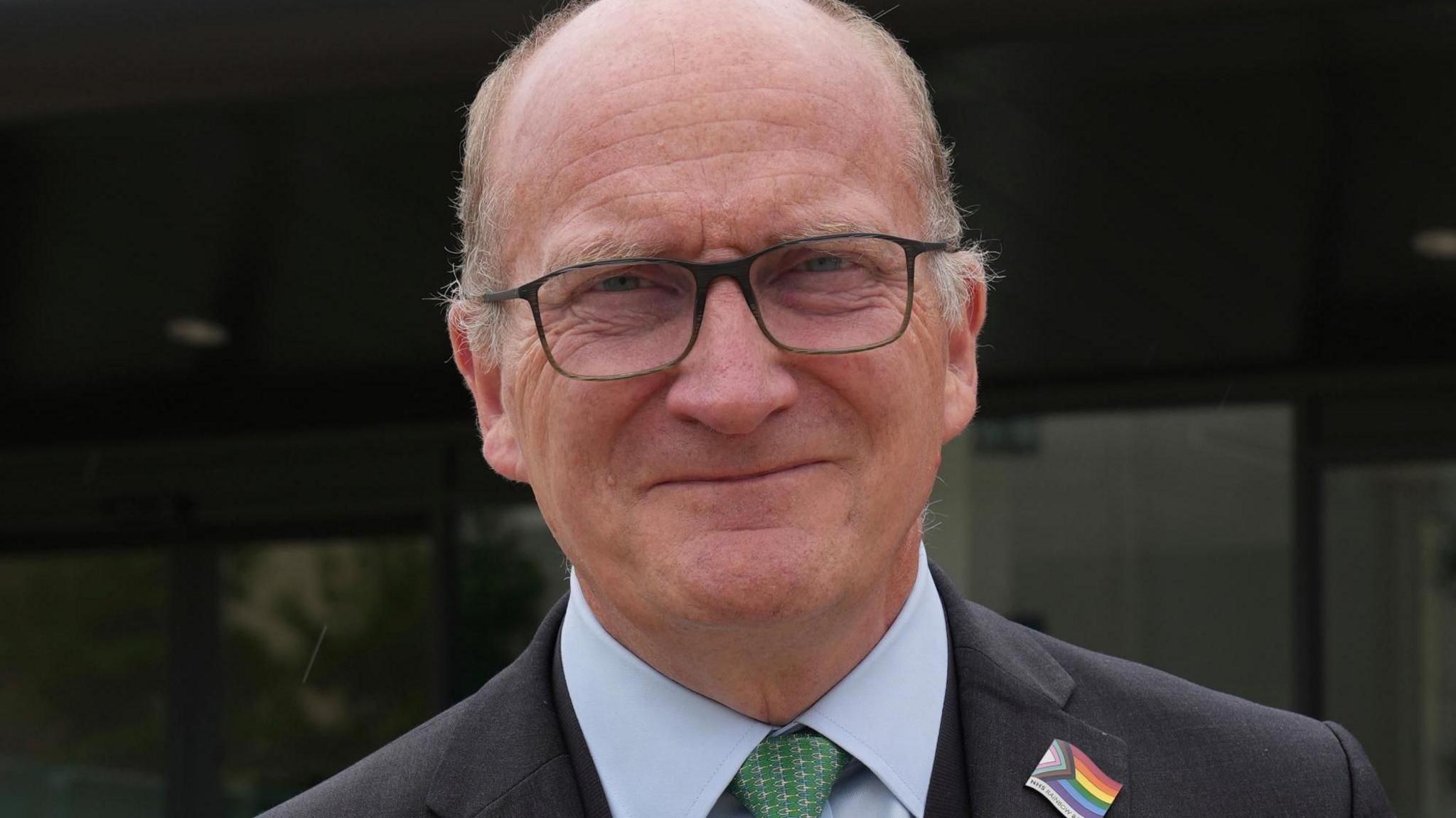 Nick Hulme is wearing a grey suit. He has a green tie on and is wearing a rainbow-coloured badge that represents LGBQT plus inclusion.