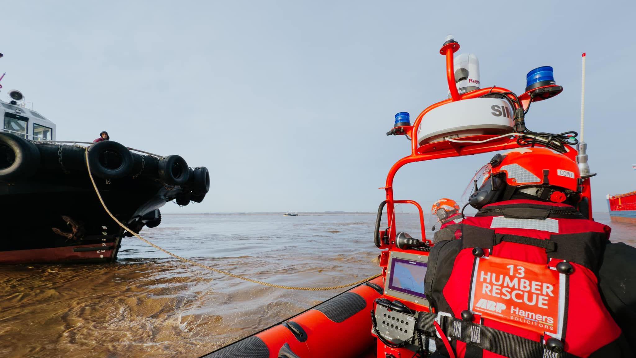 A Humber Rescue lifeboat can be seen on the right hand side out at sea with the back of one man men operating it and wearing a red lifejacket and helmet. A large vessel can be seen in the distance on the left with rope attached to it from the lifeboat.