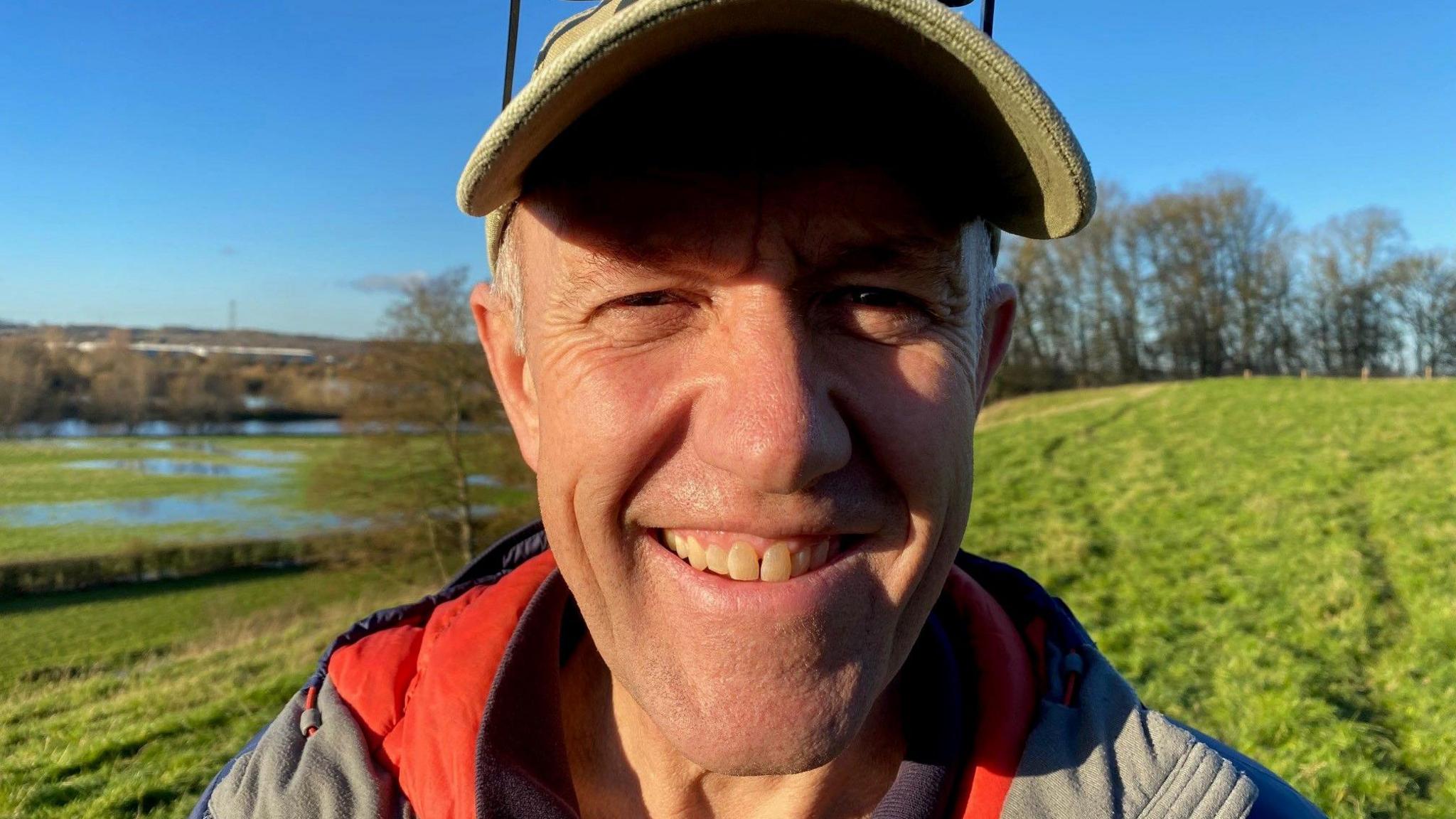 Farmer and NFU local chairman Phillip Busby standing in a field. He wears a green baseball cap, has short white hair and is clean shaven as he smiles at the camera. He wears a coat with a red collar and is in a field of green grass, next to waterway which has patches of green algae on it. Several trees are in the distance.