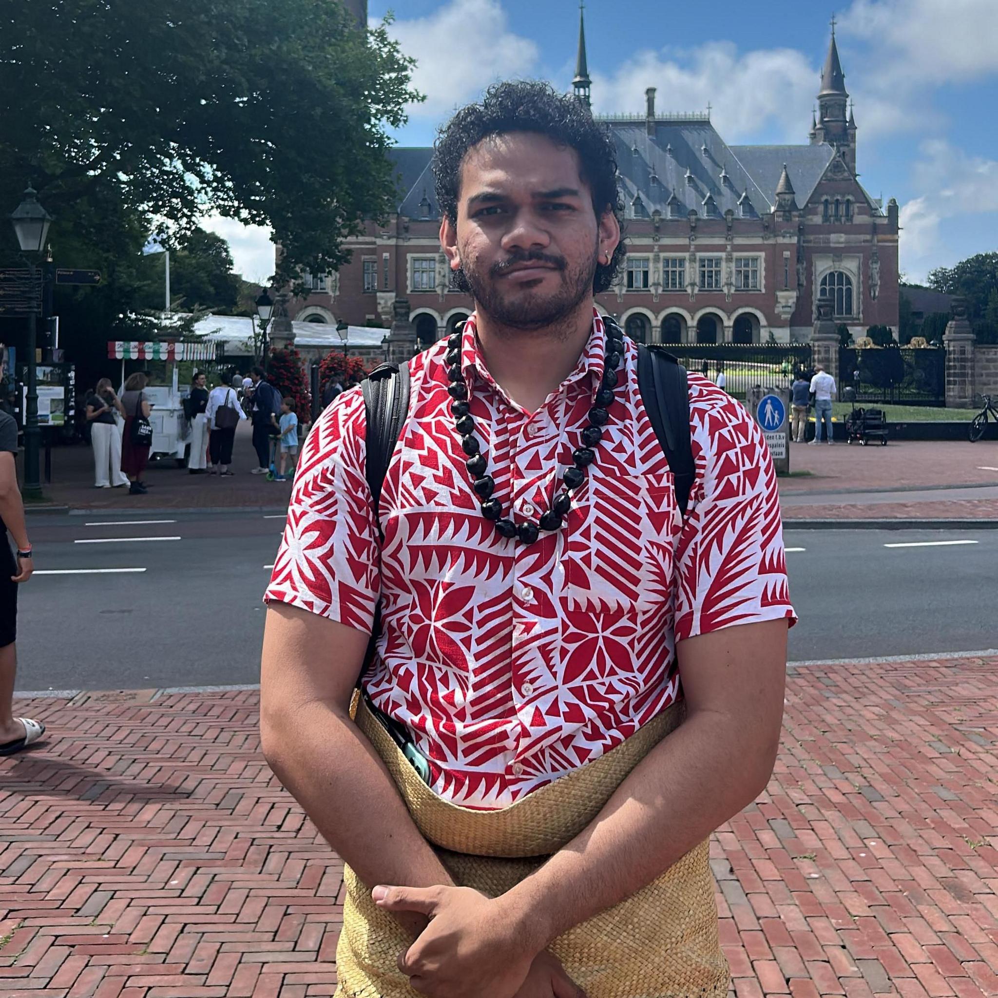 Siosiua Veikune stands with his hands clasped in front of him outside the Peace Palace in the Hague. He is wearing a red and white flower shirt and a brown necklace.