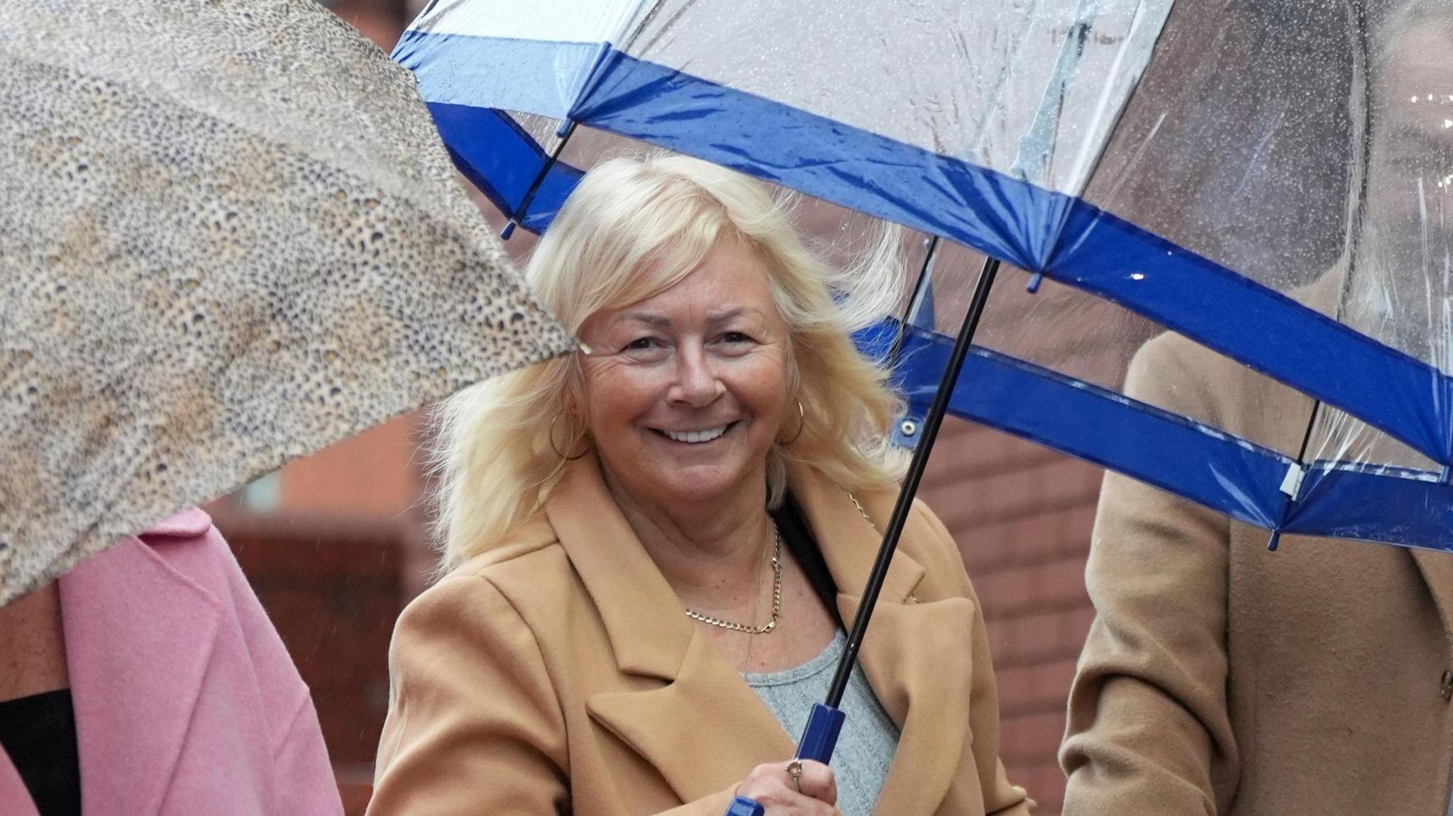 Mrs Spragg wearing a tan jacket and holding an umbrella standing next to two people either side of her