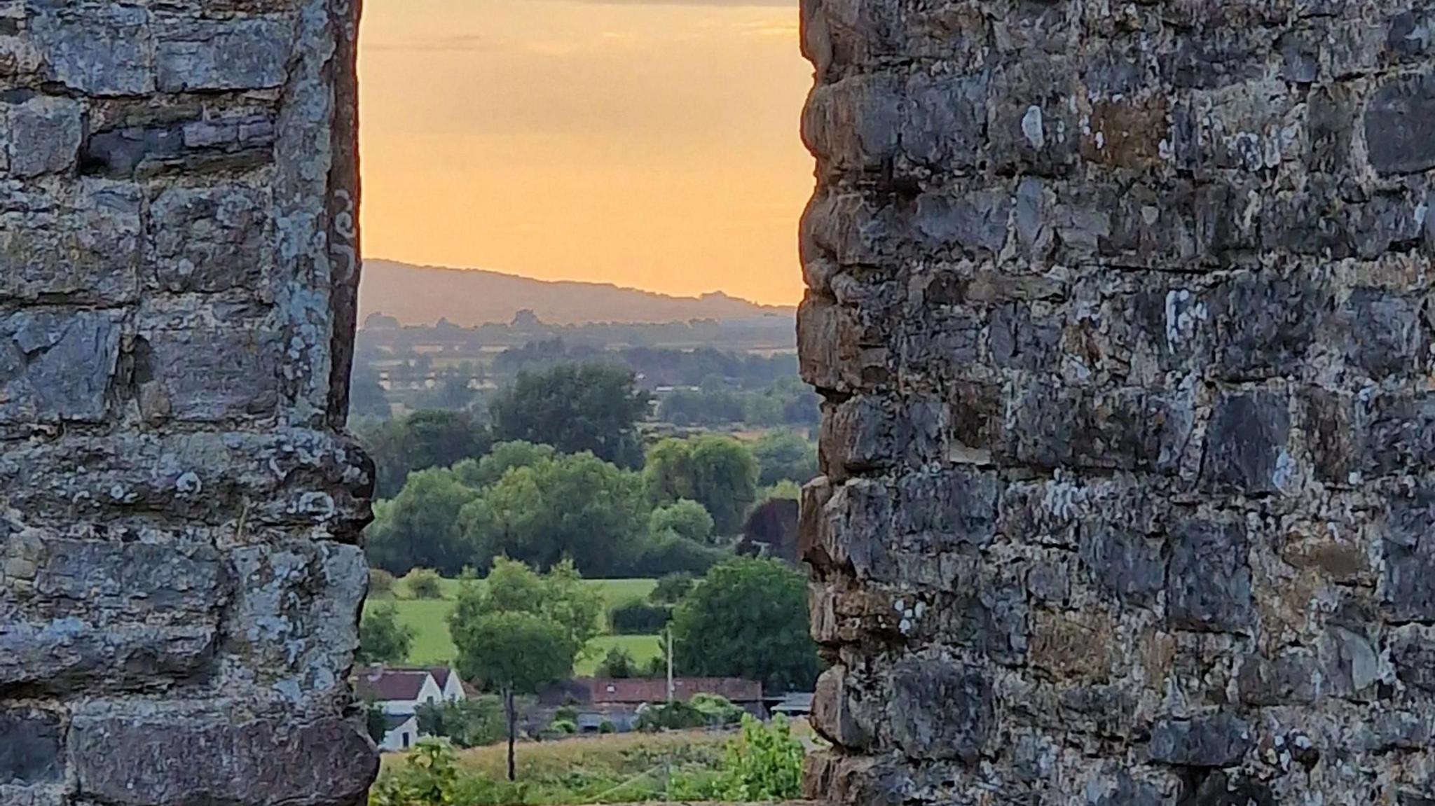 A view through a stone wall over the hills. The sun is going down colouring the sky orange.
