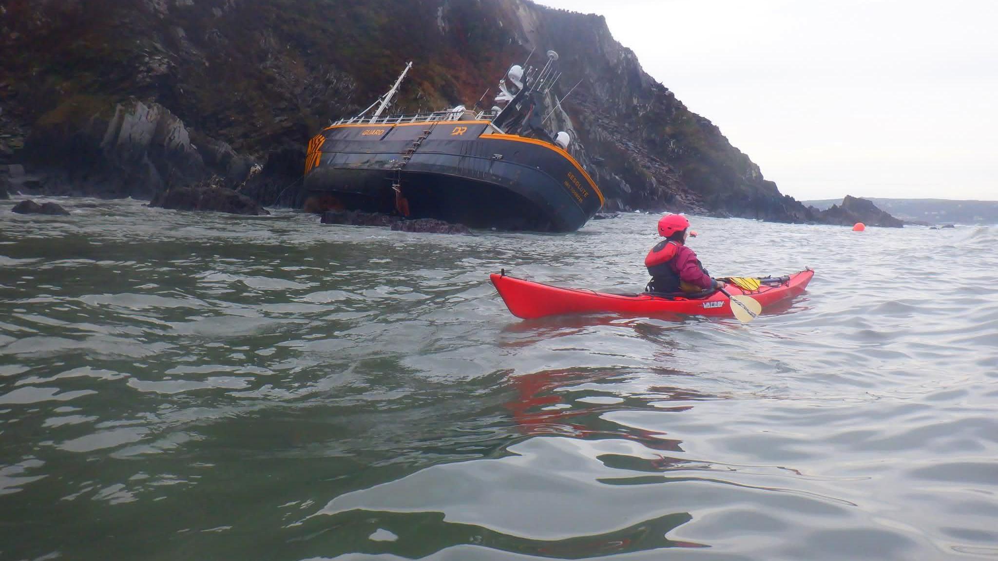 Resolute ar y creigiau yn y cefndir, yn rhannol ar ei hochr a chaiaciwr ar y môr rhwng y llong a'r camera