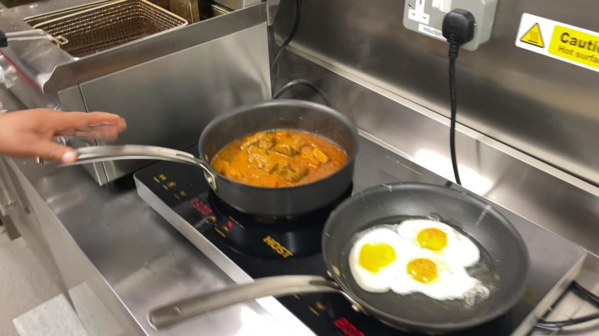 Two pans on a silver hob. One contains goat curry, the other has three fried eggs. A chef’s hand can be seen on the handle of the curry pan.