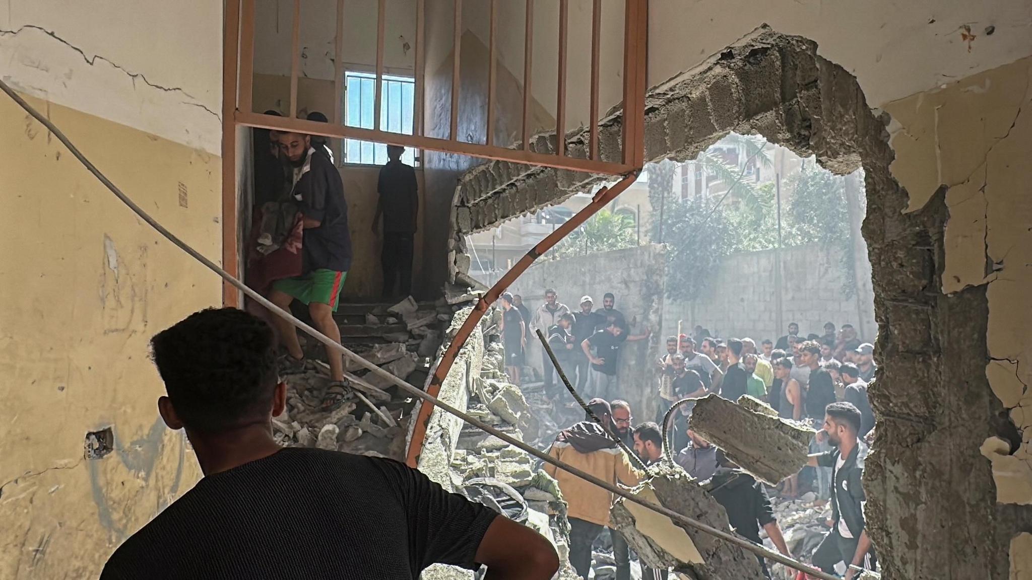 A huge hole can be seen in the side of a school building in central Gaza. Groups of people stand outside, searching through the rubble. In the top right, a man carefully carries a small body - probably a child - down a shattered staircase. At the top of the staircase, a boy looks on.