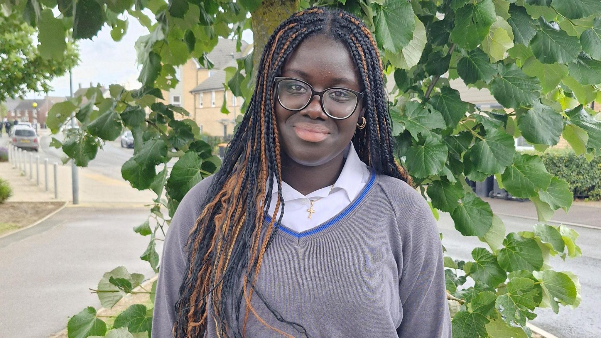 Nadine, standing outside, by a bush, smiling and looking straight at the camera, with long hair in plaits, wearing glasses, a grey school jumper and shirt.