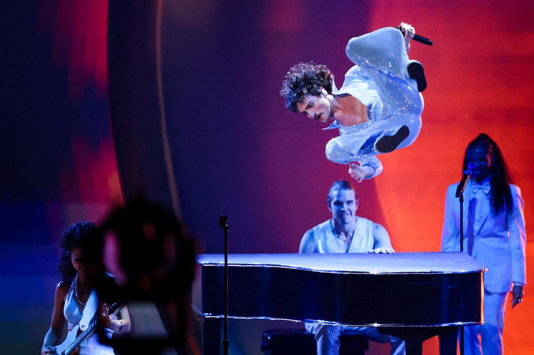 Benson Boone is pictured flying through the air during a somersault at the 67th Annual Grammy Awards in Los Angeles, California. There is a man playing a piano beneath him, a female guitarist to the left and a female singer to the right.