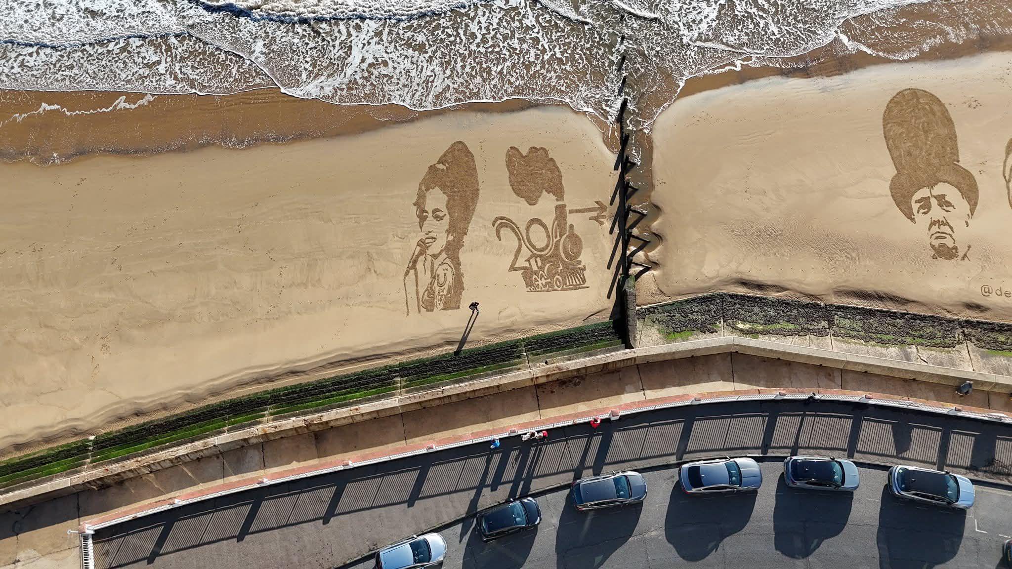 A birds-eye view of a sandy beach with Amy Winehouse, a steam train and Winston Churchill all etched onto the surface, some cars are visible on the top of the promenade.
