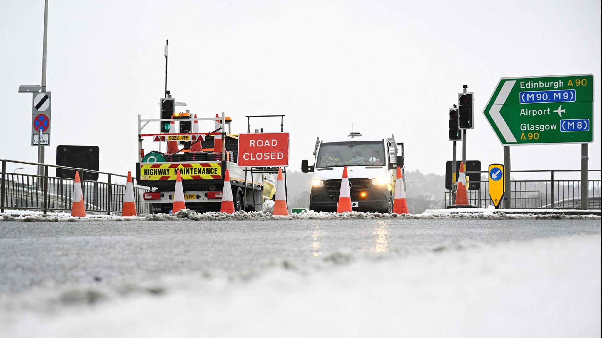 Roads vans and traffic bollards blocking an icy road with a red and white road closed sign visible