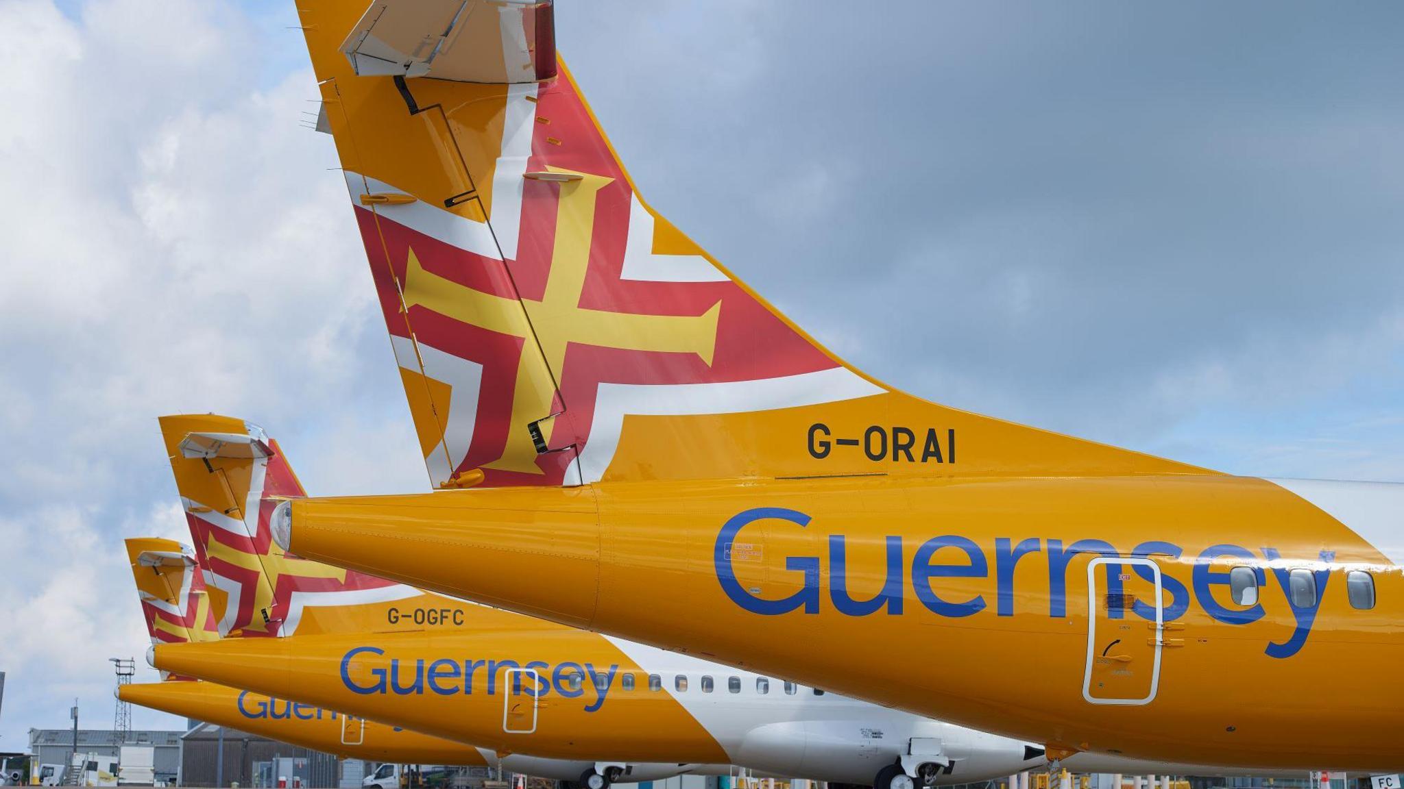 The image shows the rear of a three Aurigny planes. They are yellow with an image of the Guernsey flag on the rear fin. The Guernsey flag is a red Saint George's Cross with an additional gold Norman cross within it. The plane also has the word Guernsey written along the side in blue.