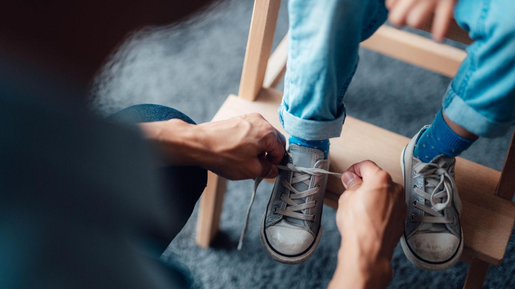 Stock close-up image showing a child's shoe laces being fastened by an adult. The youngsters is wearing blue trainers with white top caps and blue jeans.