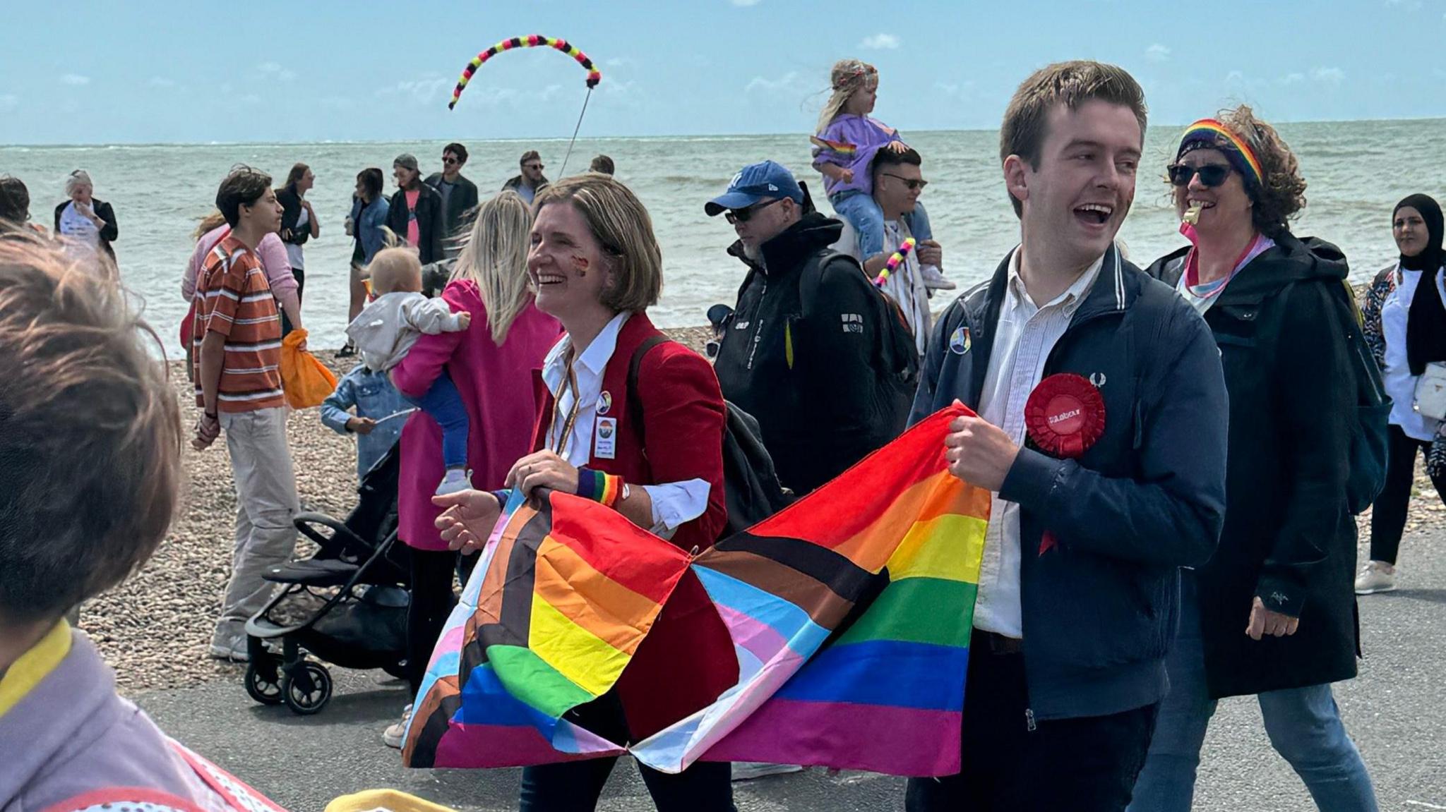 Tom Rutland and Beccy Cooper, Worthing's new MPs, hold a rainbow banner between them as they smile and walk alongside other people taking part in Worthing Pride parade