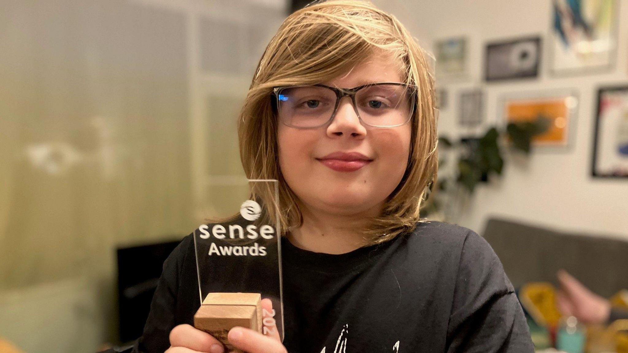 A smiling boy wearing glasses holds a trophy which says "Sense Awards" on it.