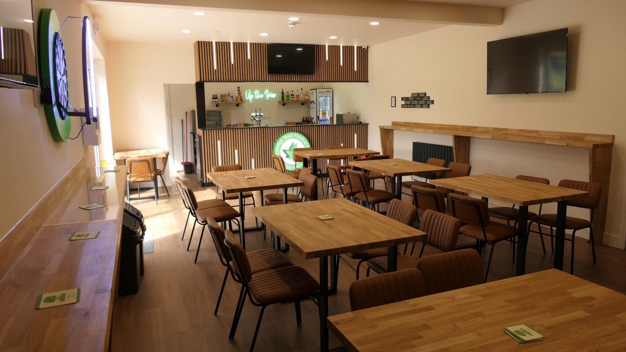 A photo of the inside of the clubhouse. It has lots of seating and wooden tables with a standing bar area. There is a dartboard and a TV on the wall. The club's logo is prominently on display in front of the bar and lit up.